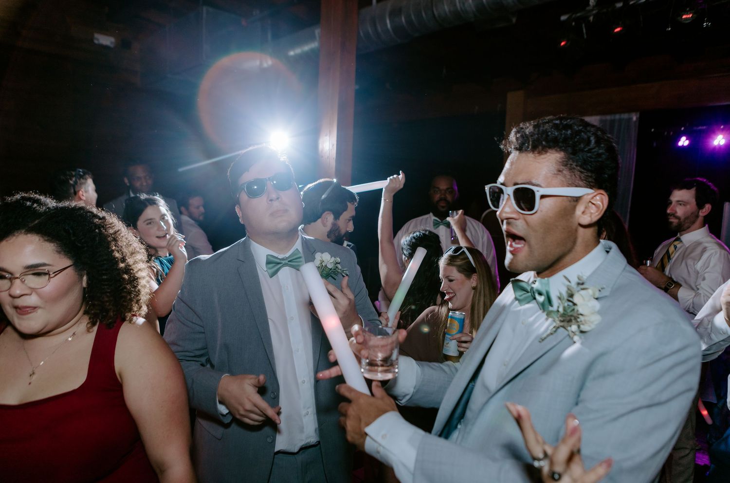 Wedding guests dancing at reception, two men in suits wearing white sunglasses celebrating on dance floor.