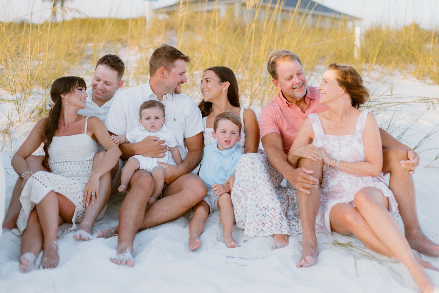 A joyful group sits together on white sand beach at sunset with coastal grasses in the background.