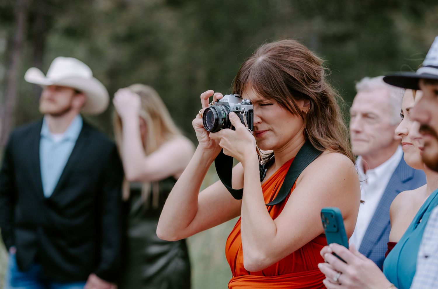 Woman in orange dress photographs wedding guests outdoors with a film camera.