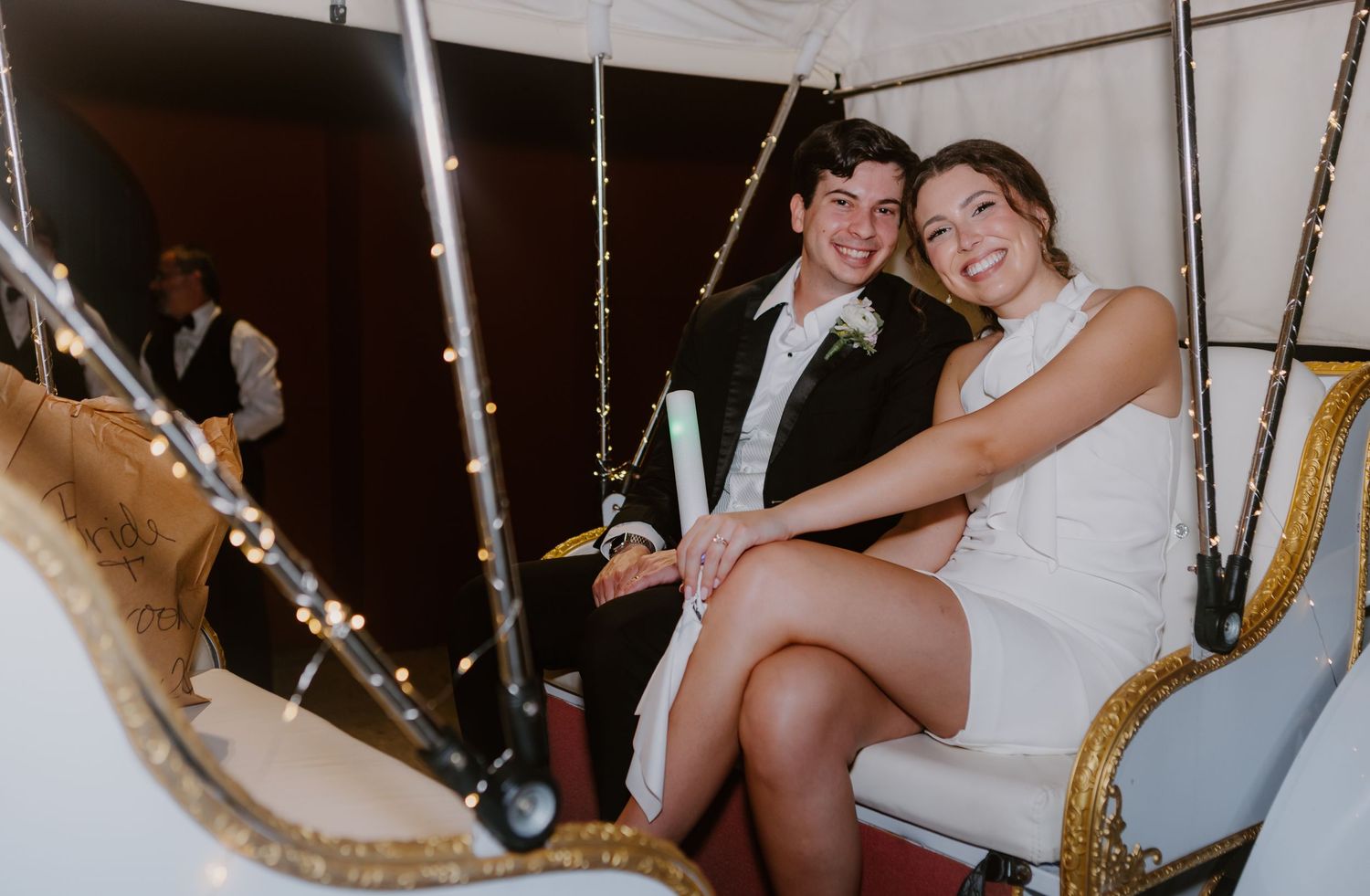 Newlyweds sit together smiling inside a decorated white horse-drawn carriage at night.