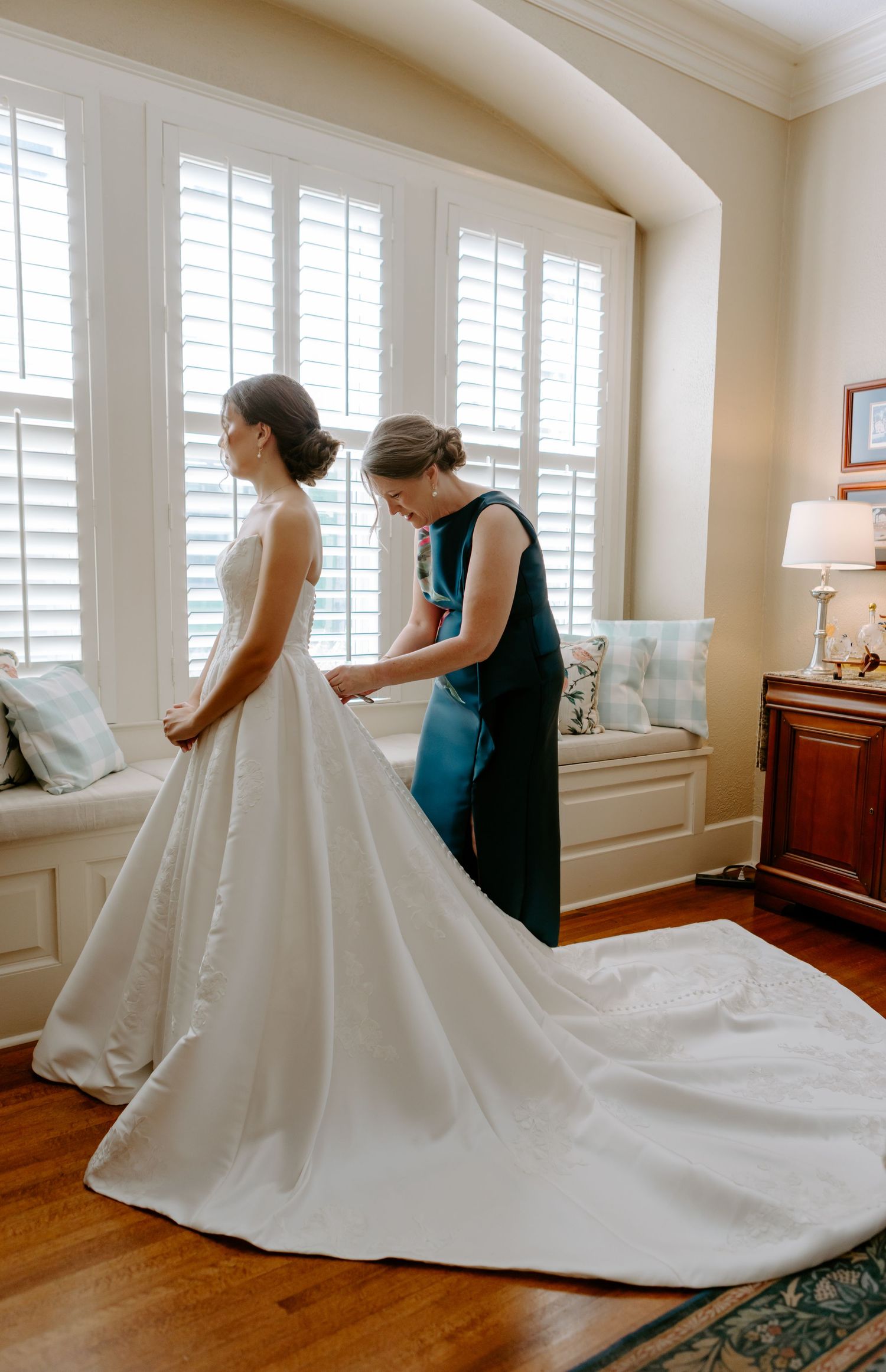A bride in a white ball gown gets her dress fastened by a woman in teal near bright plantation shuttered windows.