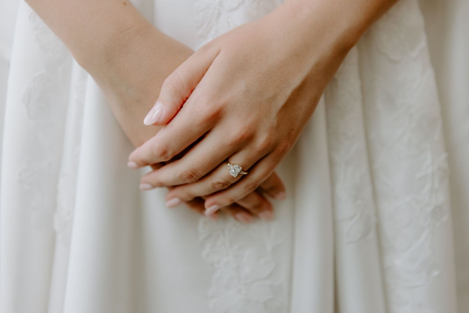 Close-up of bride's hands clasped together wearing an elegant diamond engagement ring on white wedding dress.