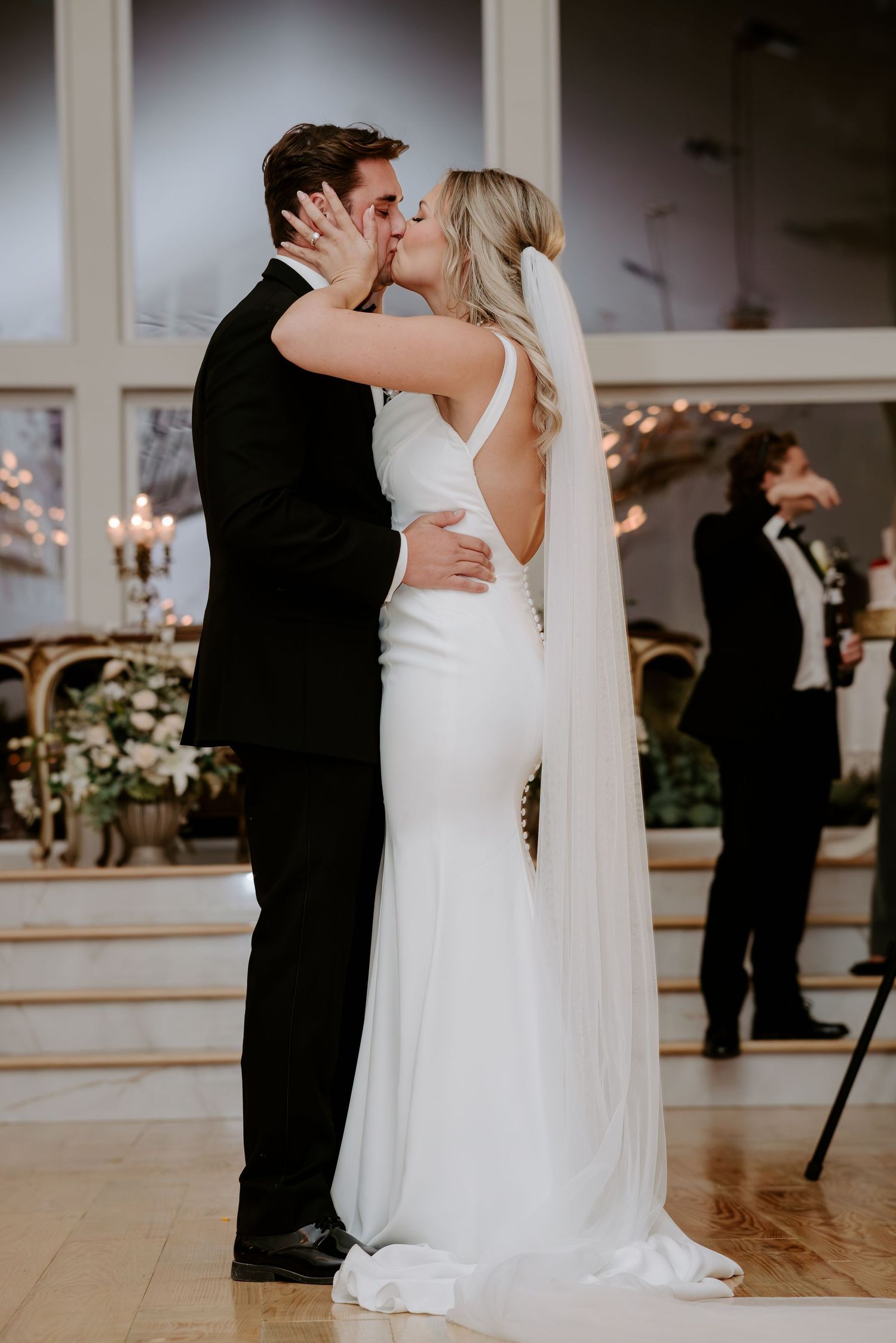 Bride in a white backless gown with a long veil kisses her groom in a black tuxedo during a wedding ceremony in an elegant venue.