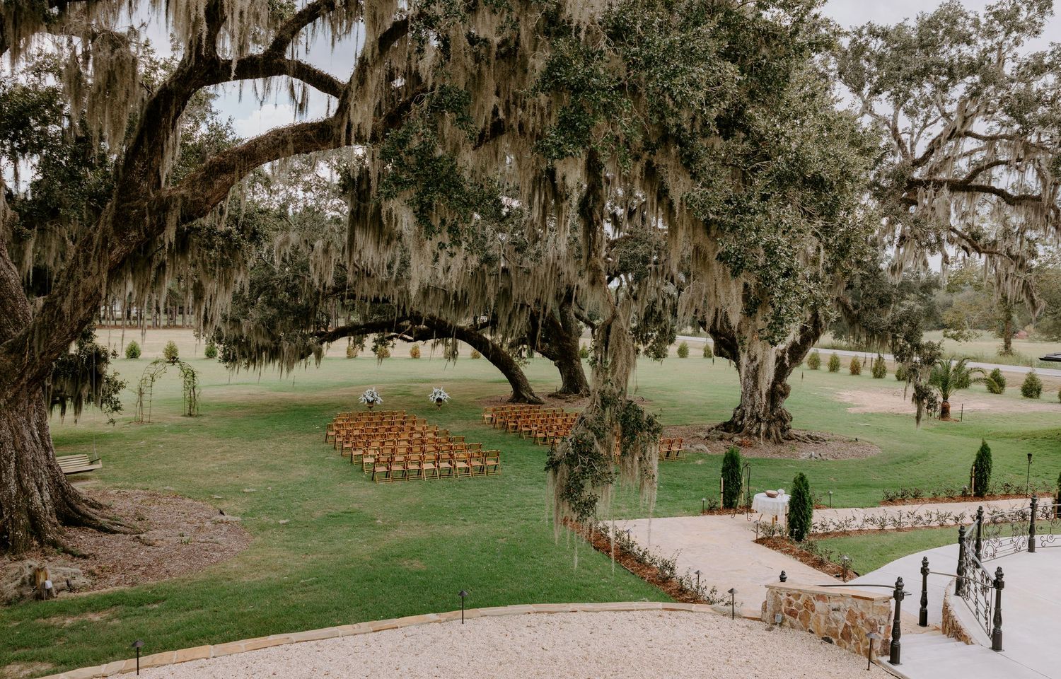 Outdoor wedding ceremony setup with rows of wooden chairs on a green lawn beneath live oak trees draped in Spanish moss.