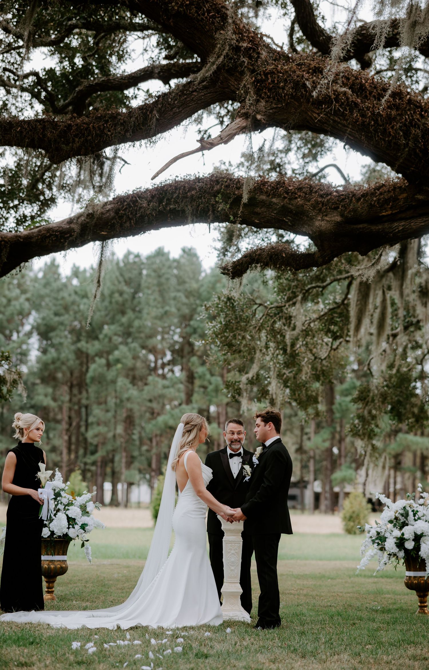 Bride and groom hold hands during their outdoor wedding ceremony under a moss-draped oak tree as a bridesmaid watches nearby.