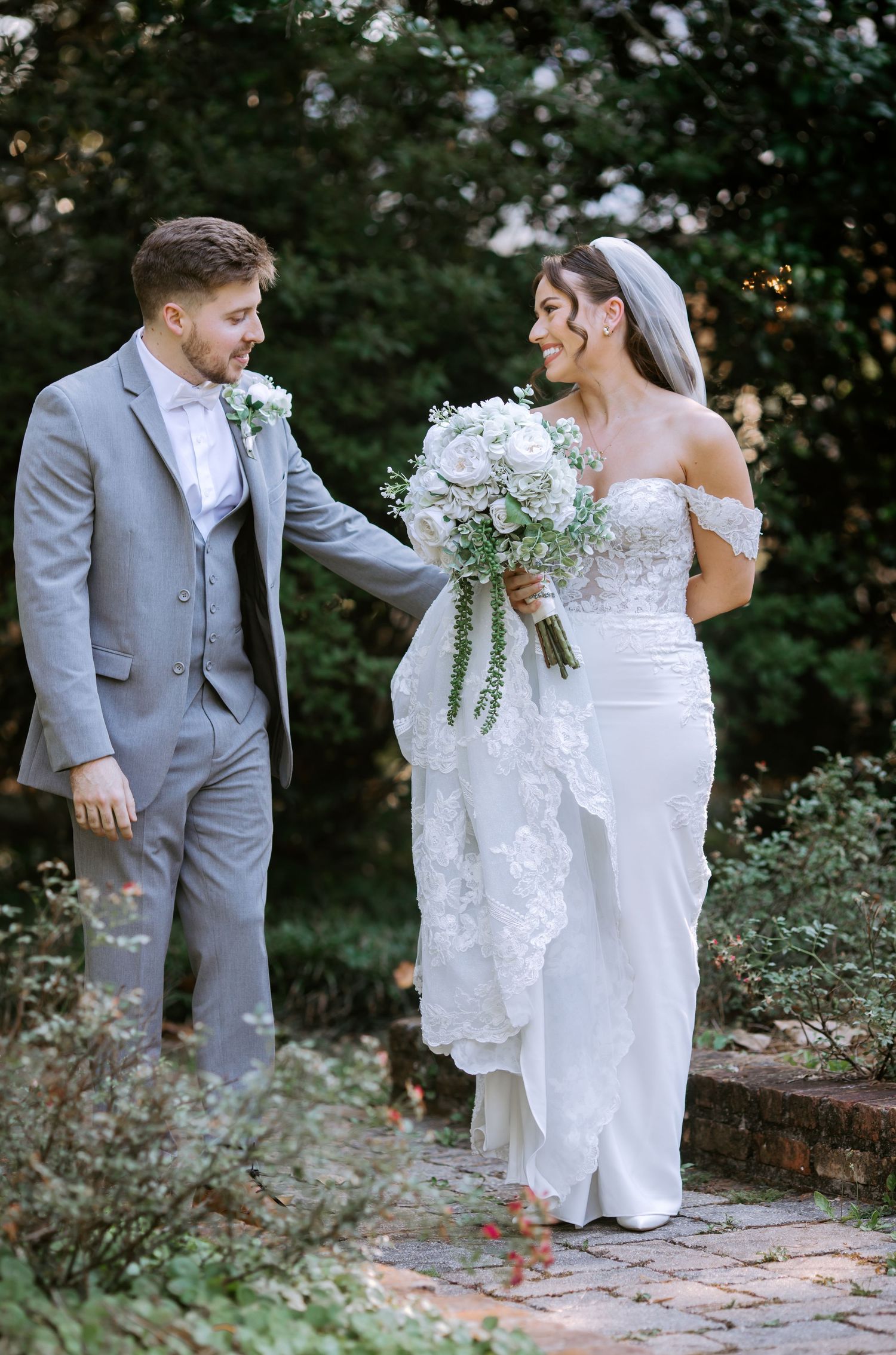 Bride and groom walking hand in hand in a lush garden, bride holding white floral bouquet in elegant lace wedding gown.