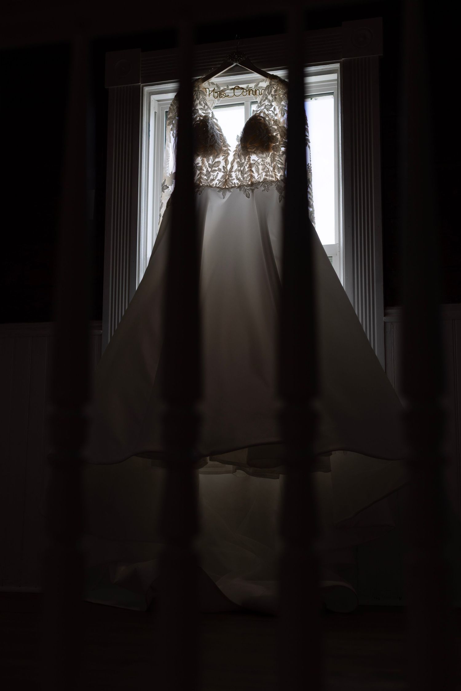 Elegant wedding dress hanging in a window, featuring lace bodice and flowing skirt, backlit by natural light.