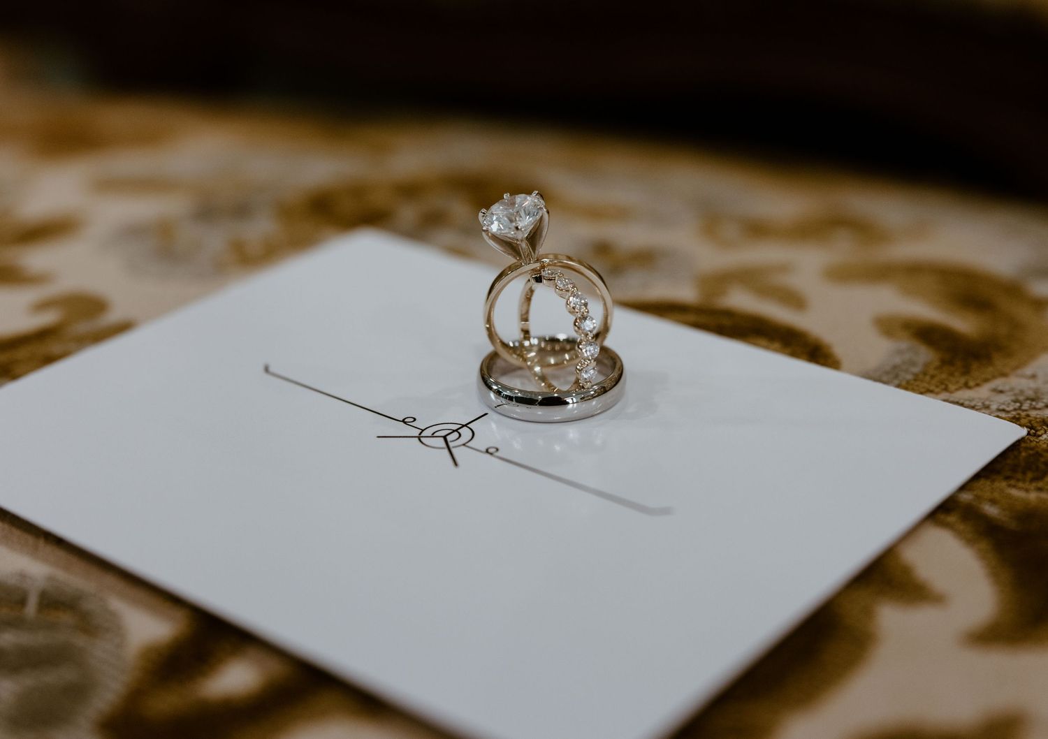 Diamond solitaire engagement ring stacked on a wedding band, resting on a white invitation card atop an ornate gold patterned fabric.