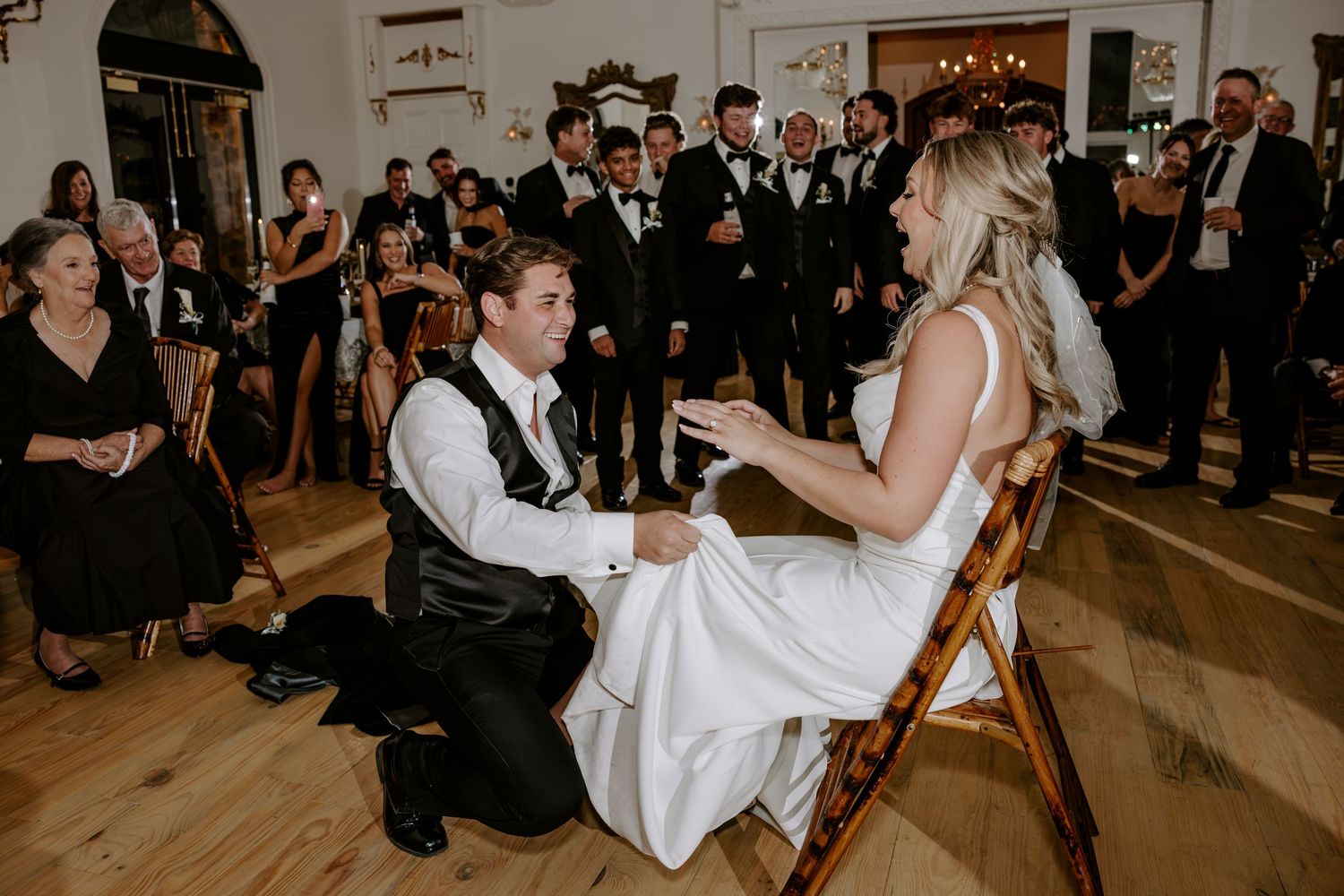Groom kneels on a wooden dance floor removing the garter from a seated bride in a white gown as wedding guests in formal attire watch.