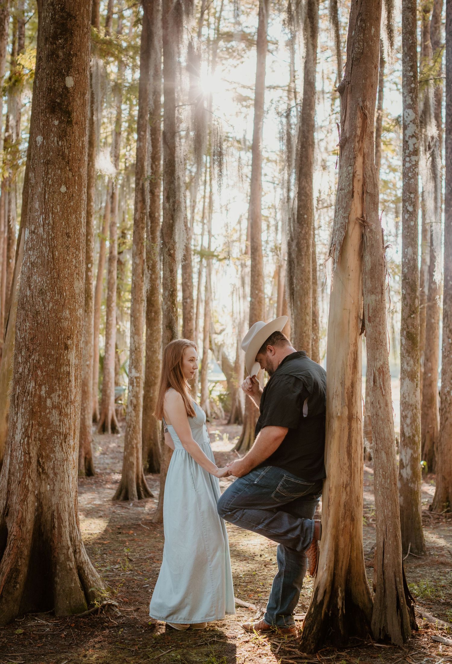 A couple shares a tender moment among tall trees in a sunlit forest setting during a romantic photoshoot.