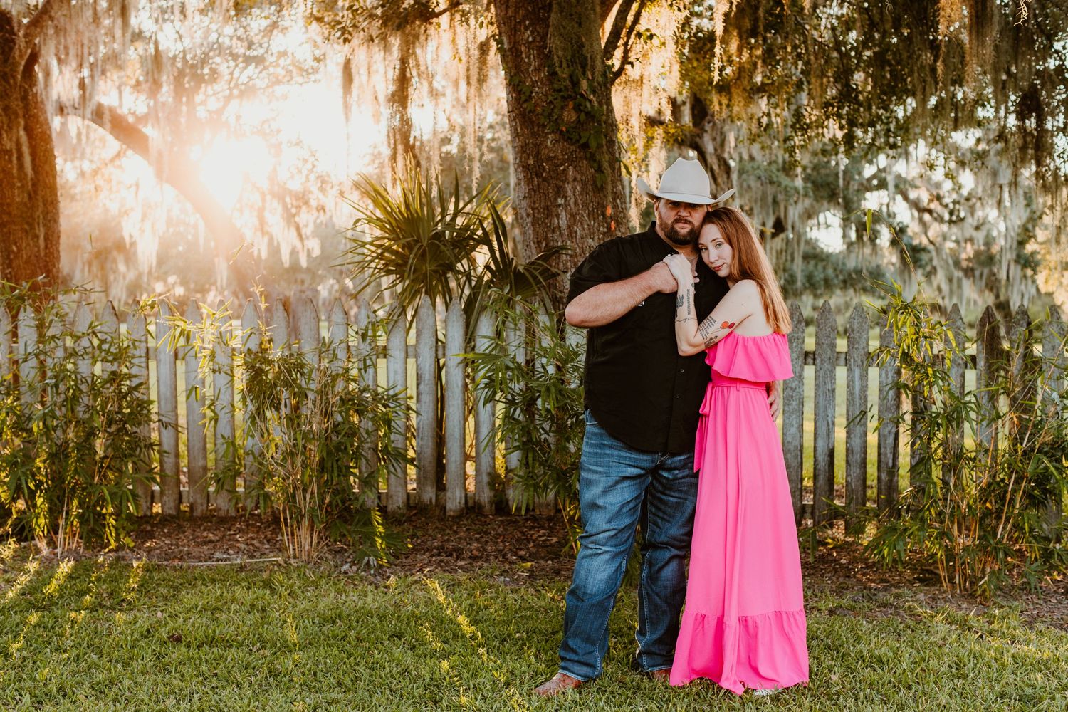 A couple embraces in a backyard garden setting with sunlight streaming through trees in the background.
