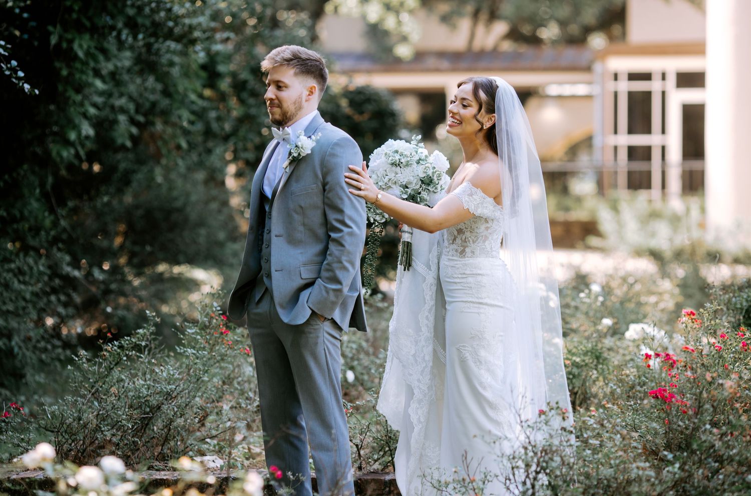 Bride in lace gown and veil holds bouquet behind groom in gray suit during outdoor garden wedding ceremony.