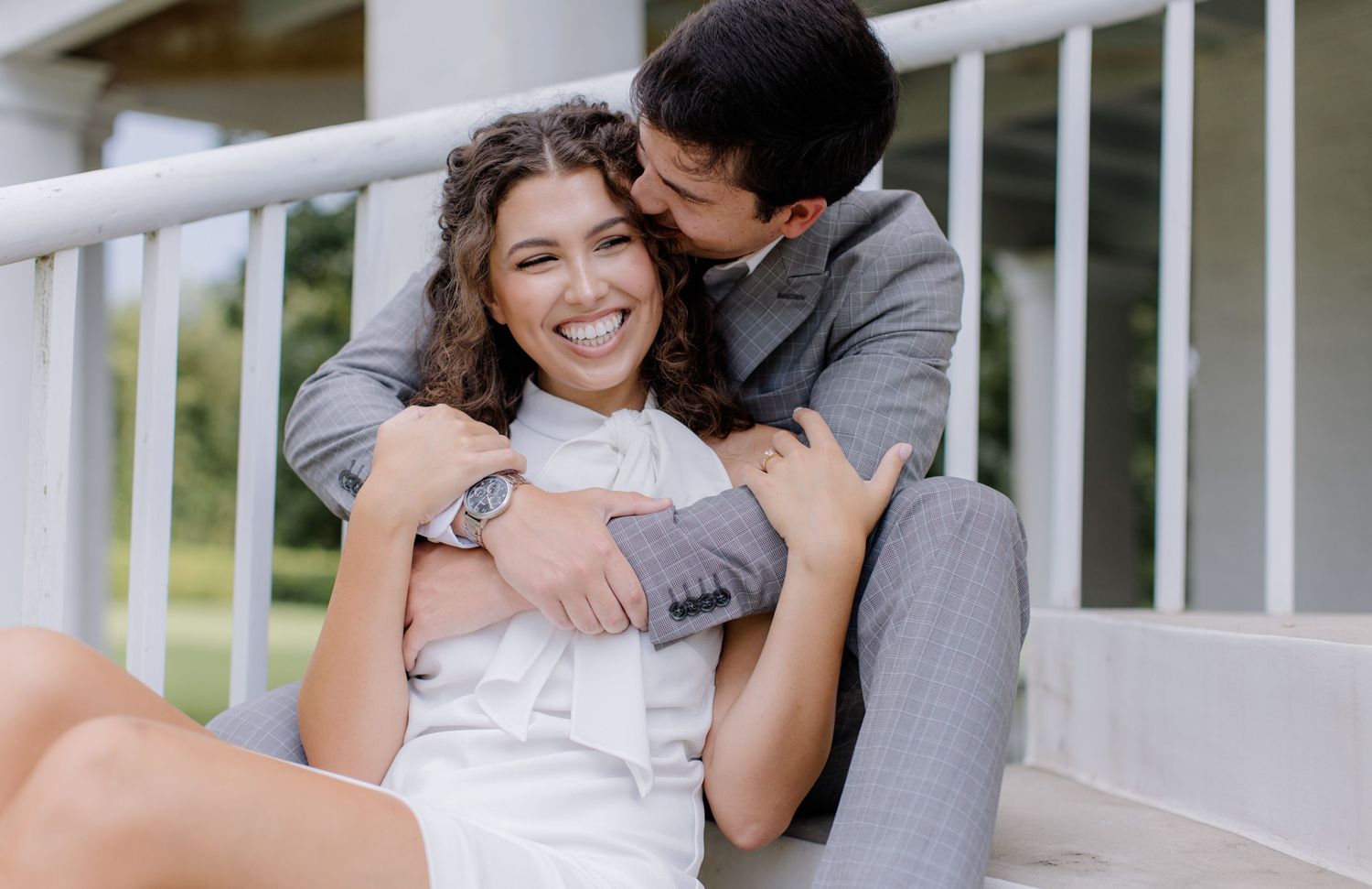A couple in formal wedding attire share an intimate moment on a white porch railing outdoors.