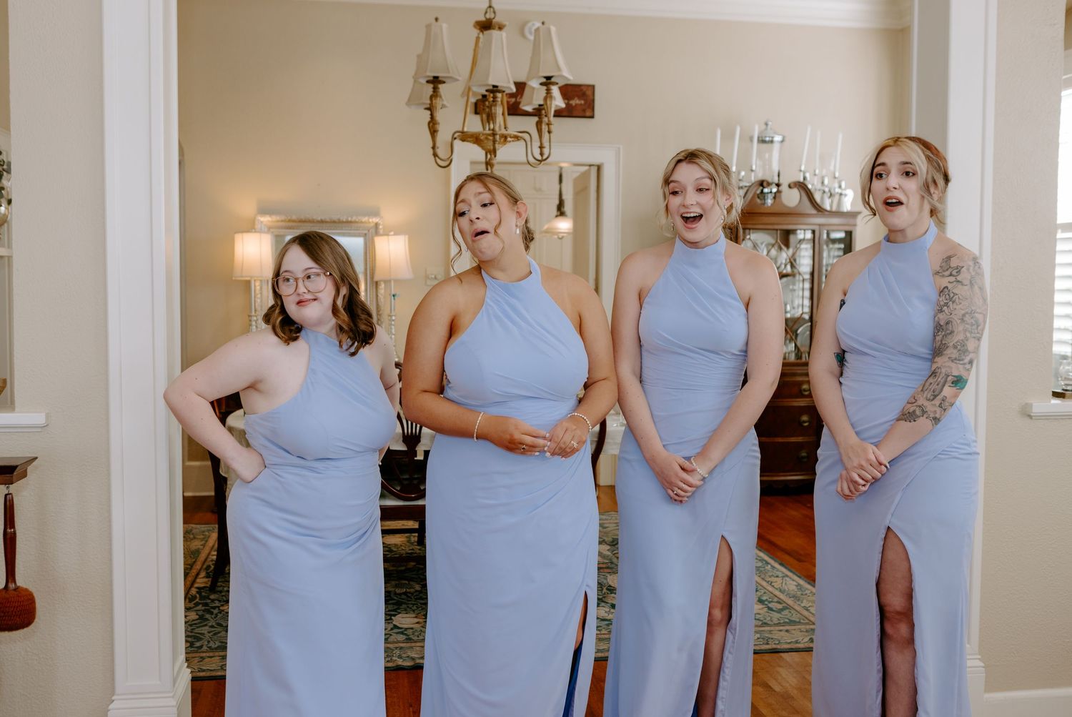 Four bridesmaids in light blue halter gowns reacting with excitement indoors.
