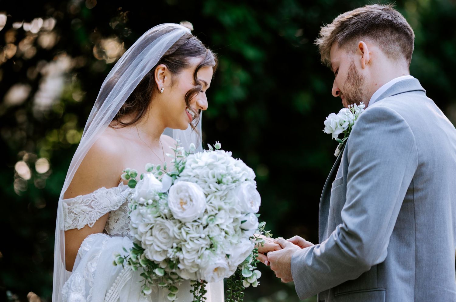 Bride and groom exchanging rings outdoors, bride holding white floral bouquet wearing lace off-shoulder gown and veil.