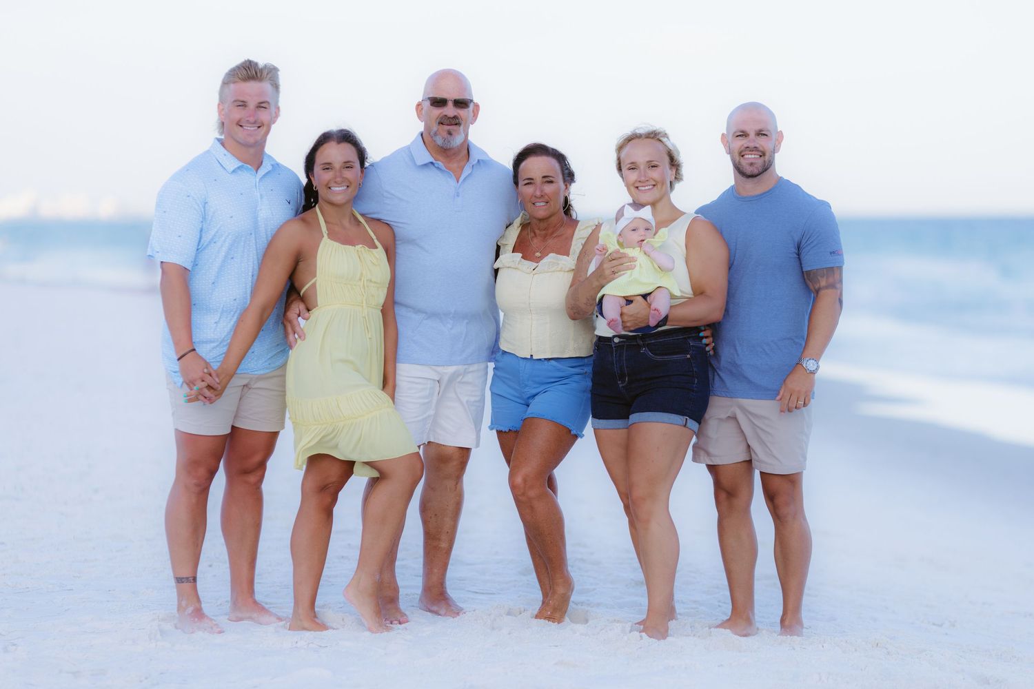 Multi-generational family wearing casual summer clothes poses together on a sunny beach with ocean waves in background.