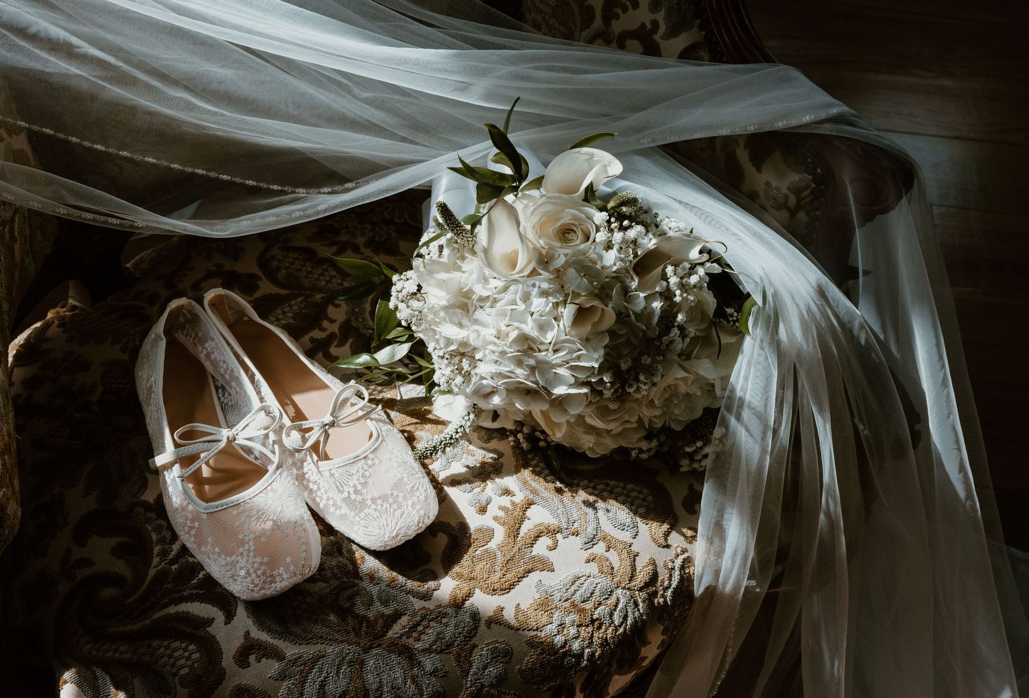 Lace bridal flats and a white rose bouquet rest on an ornate patterned chair beneath a sheer wedding veil in soft window light.