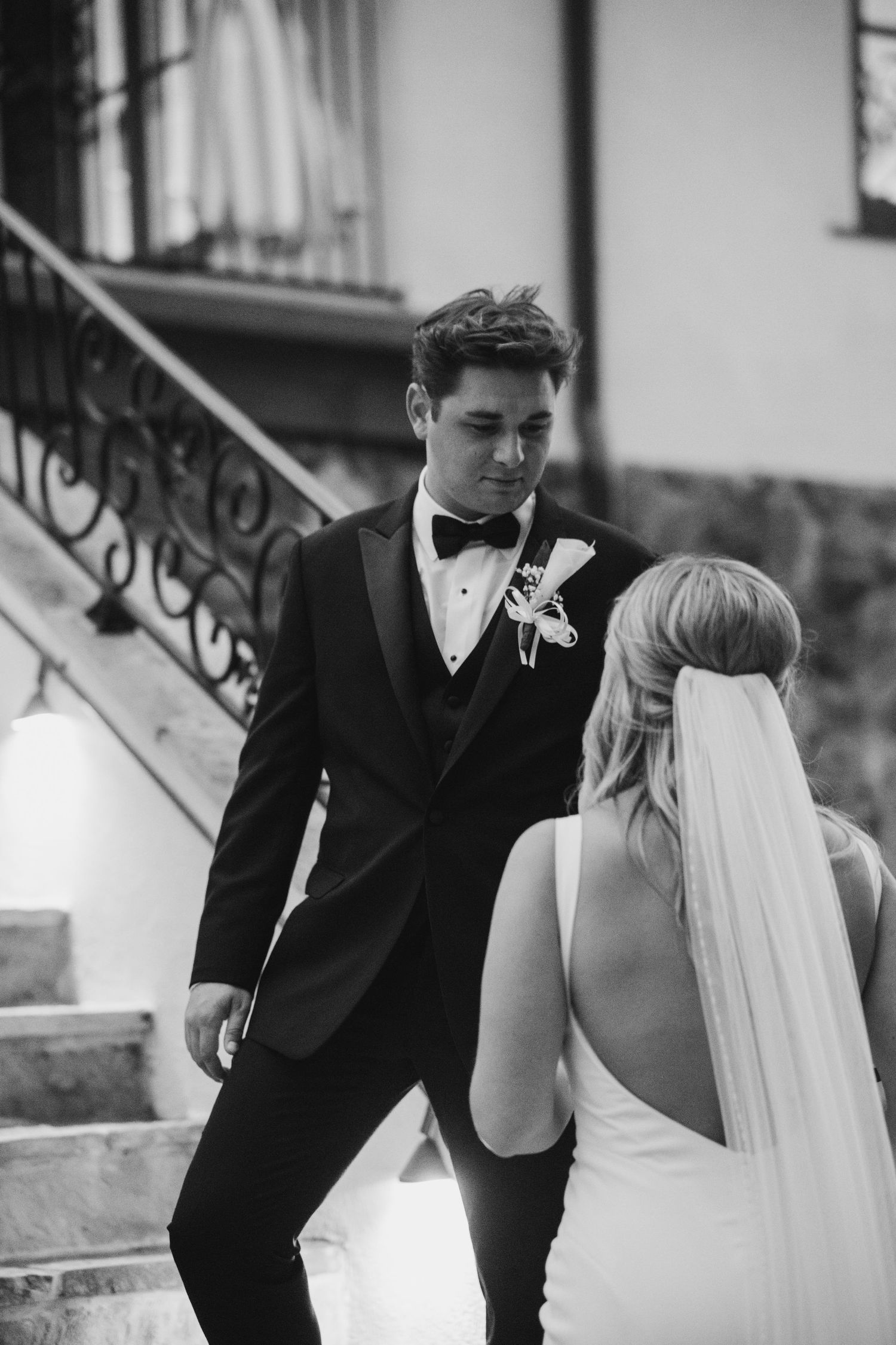 Black and white photo of a groom in a tuxedo and bow tie looking down at a bride in a veil and backless gown beside an ornate staircase.