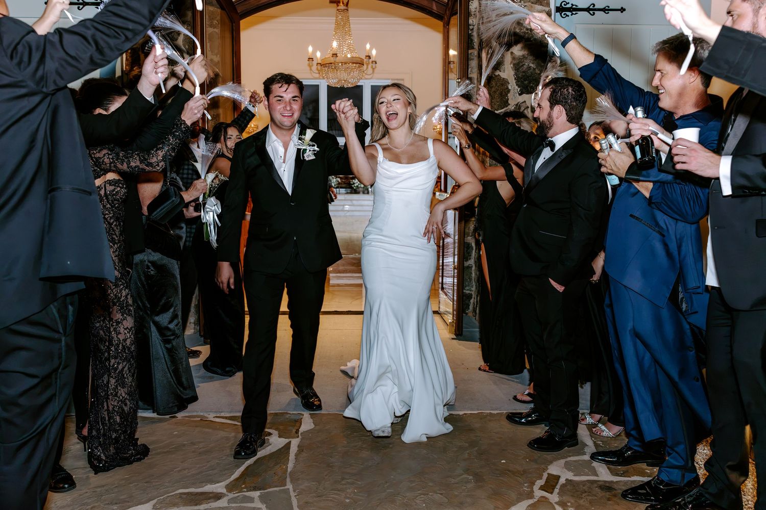 Bride in a white gown and groom in a dark suit walk hand in hand through guests holding sparklers at a wedding exit.