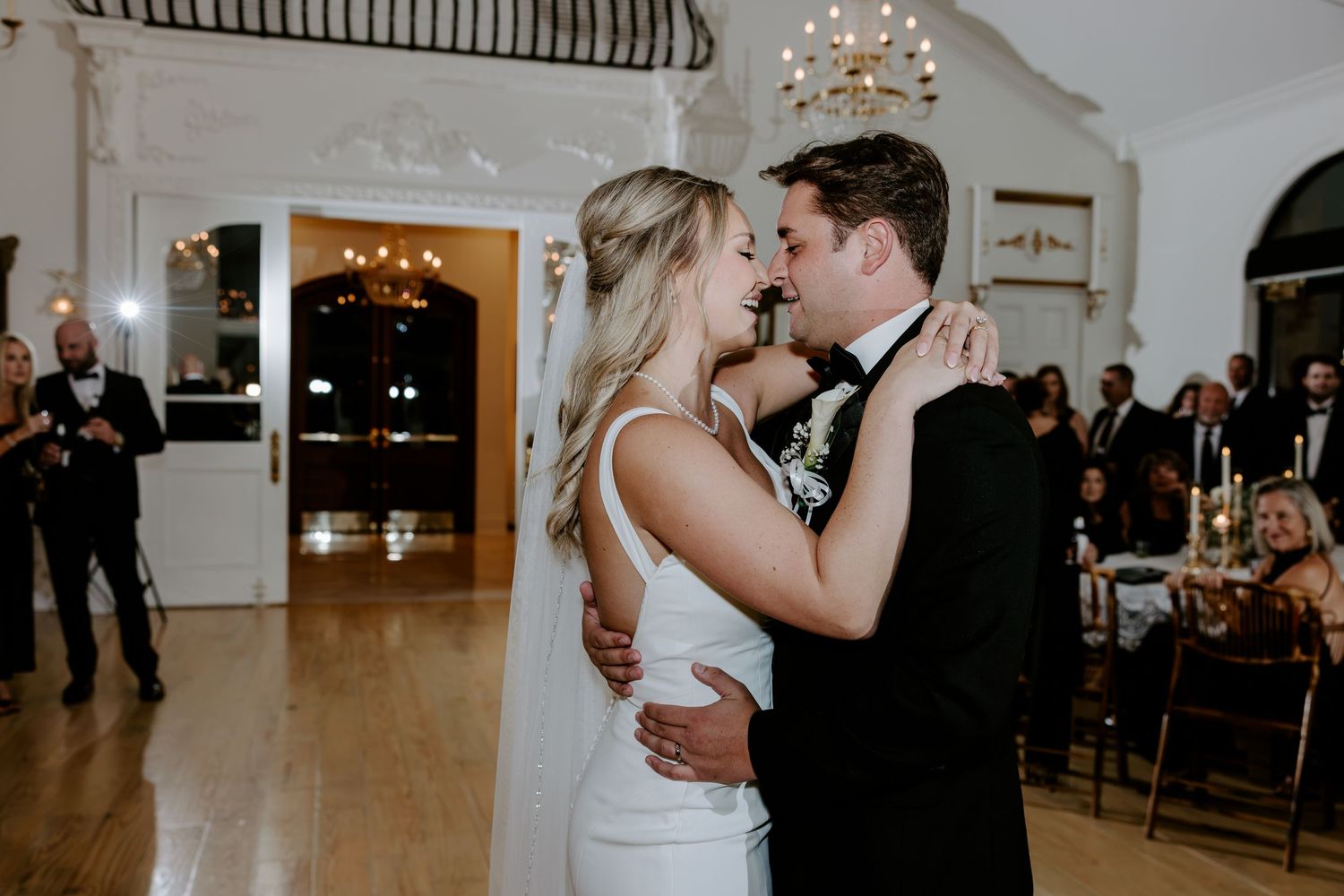 Bride in a white gown and groom in a black tuxedo share a first dance as wedding guests watch in an elegant ballroom.