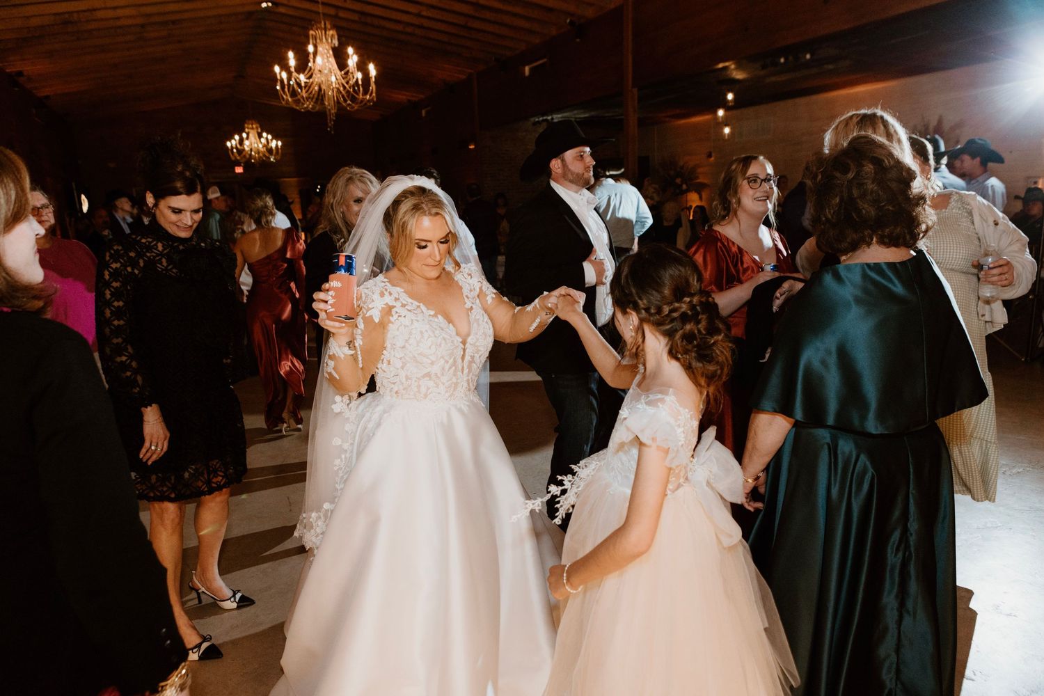 Bride in white ball gown dancing with flower girl at elegant wedding reception in rustic venue.