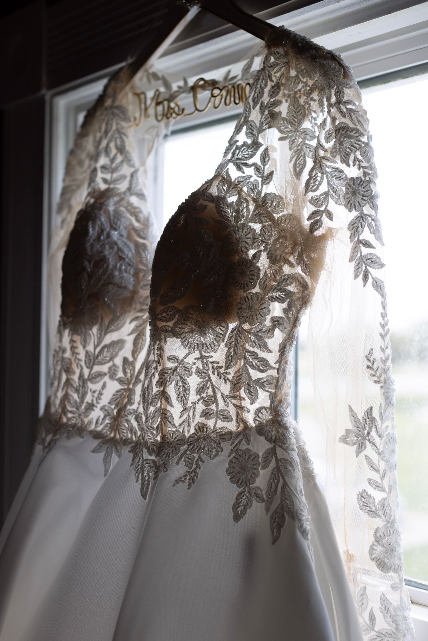 Elegant wedding dress with intricate gold lace embroidery hanging by a window on a personalized bridal hanger.