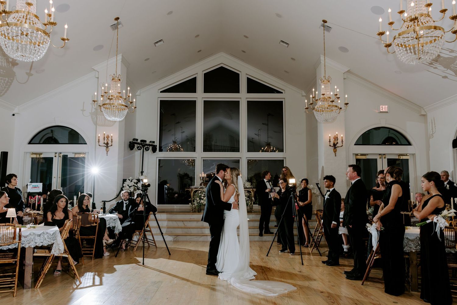 Bride and groom share a first dance in an elegant ballroom with chandeliers, tall windows, and wedding guests and photographers watching.