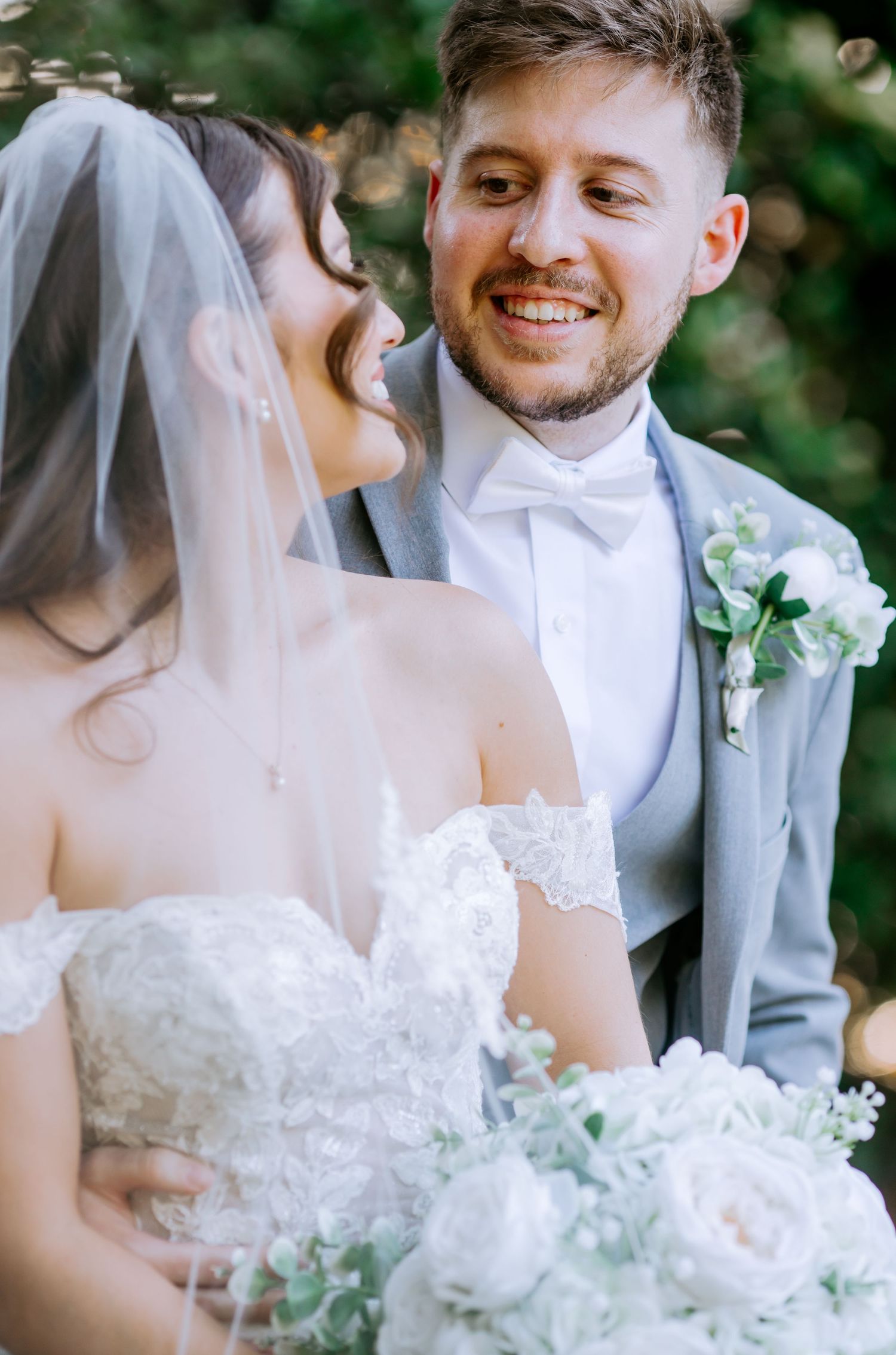 Bride and groom share a joyful moment, she in lace off-shoulder dress with veil, he in gray suit with boutonniere.