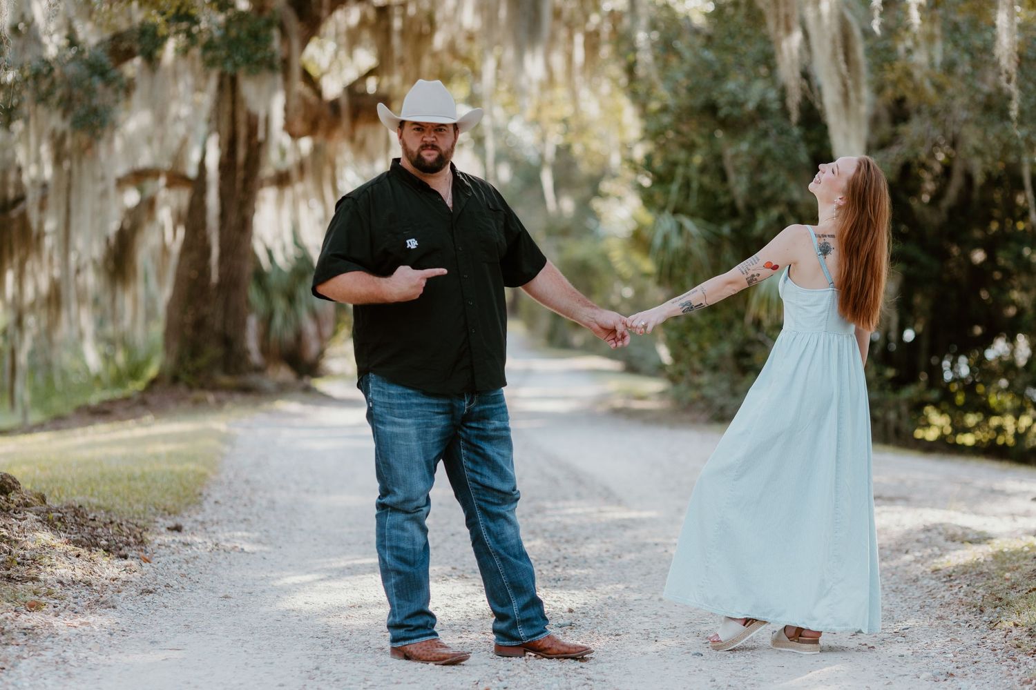 A couple in a light blue dress and casual attire hold hands while walking along a tree-lined path.