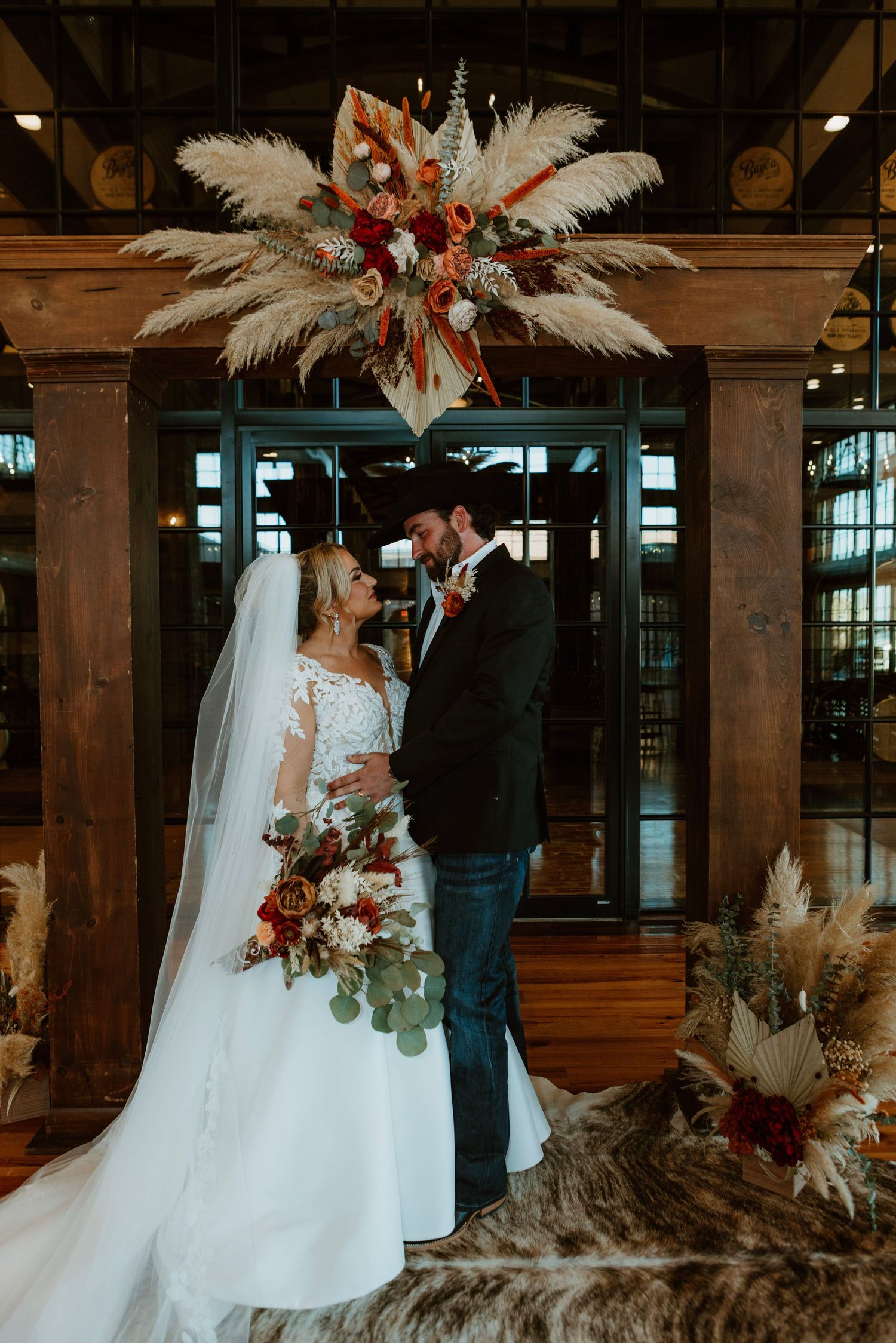 Bride and groom stand under boho pampas grass floral arch in rustic venue, bride holding burgundy bouquet.