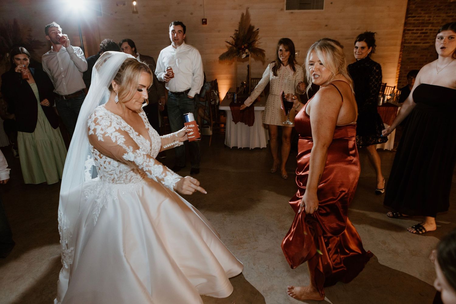 Bride in white gown dancing with guest in orange dress at wedding reception celebration.
