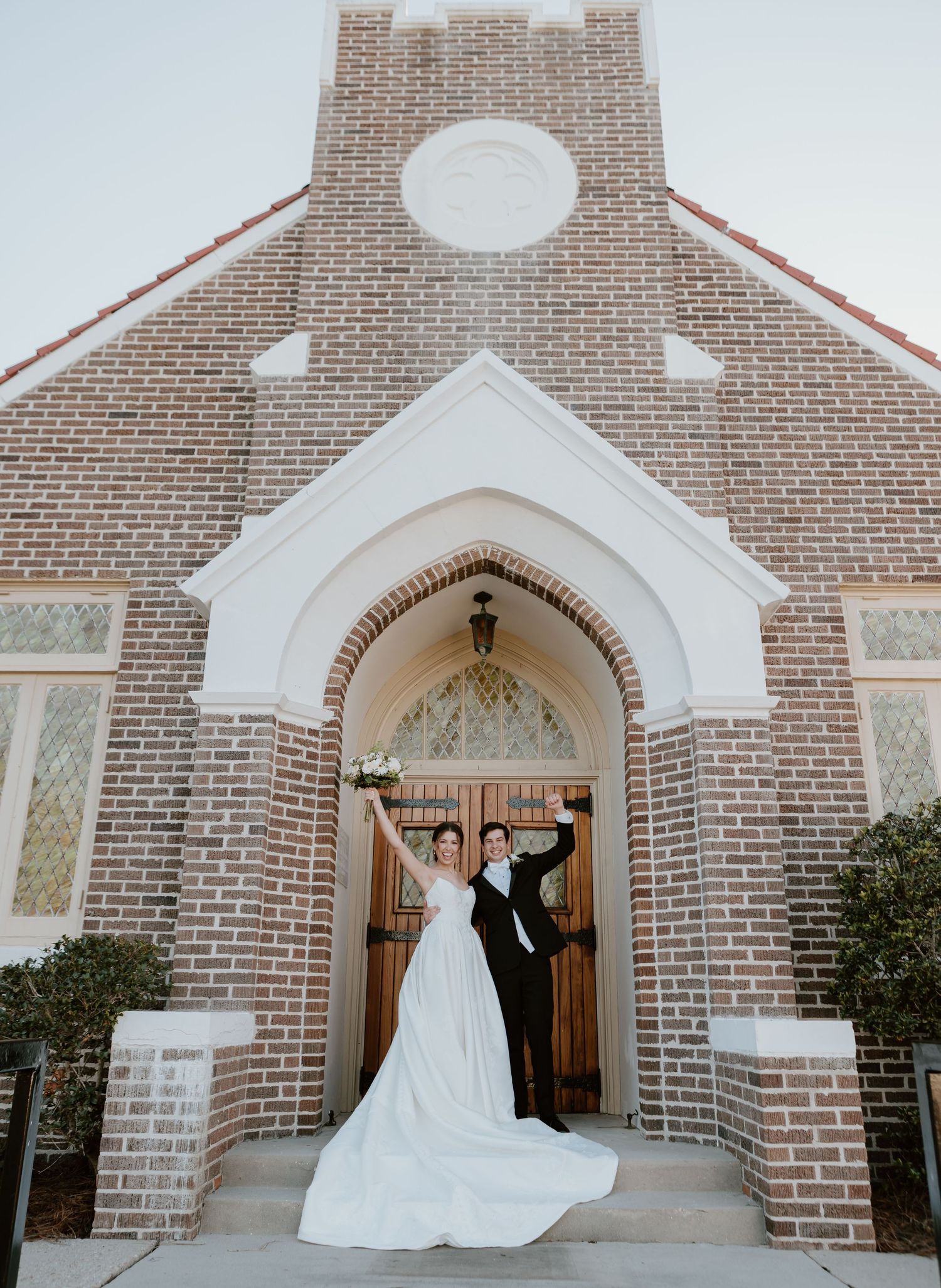 Newlywed couple celebrating outside a brick church wedding venue, bride in white gown and groom in black suit raising hands.