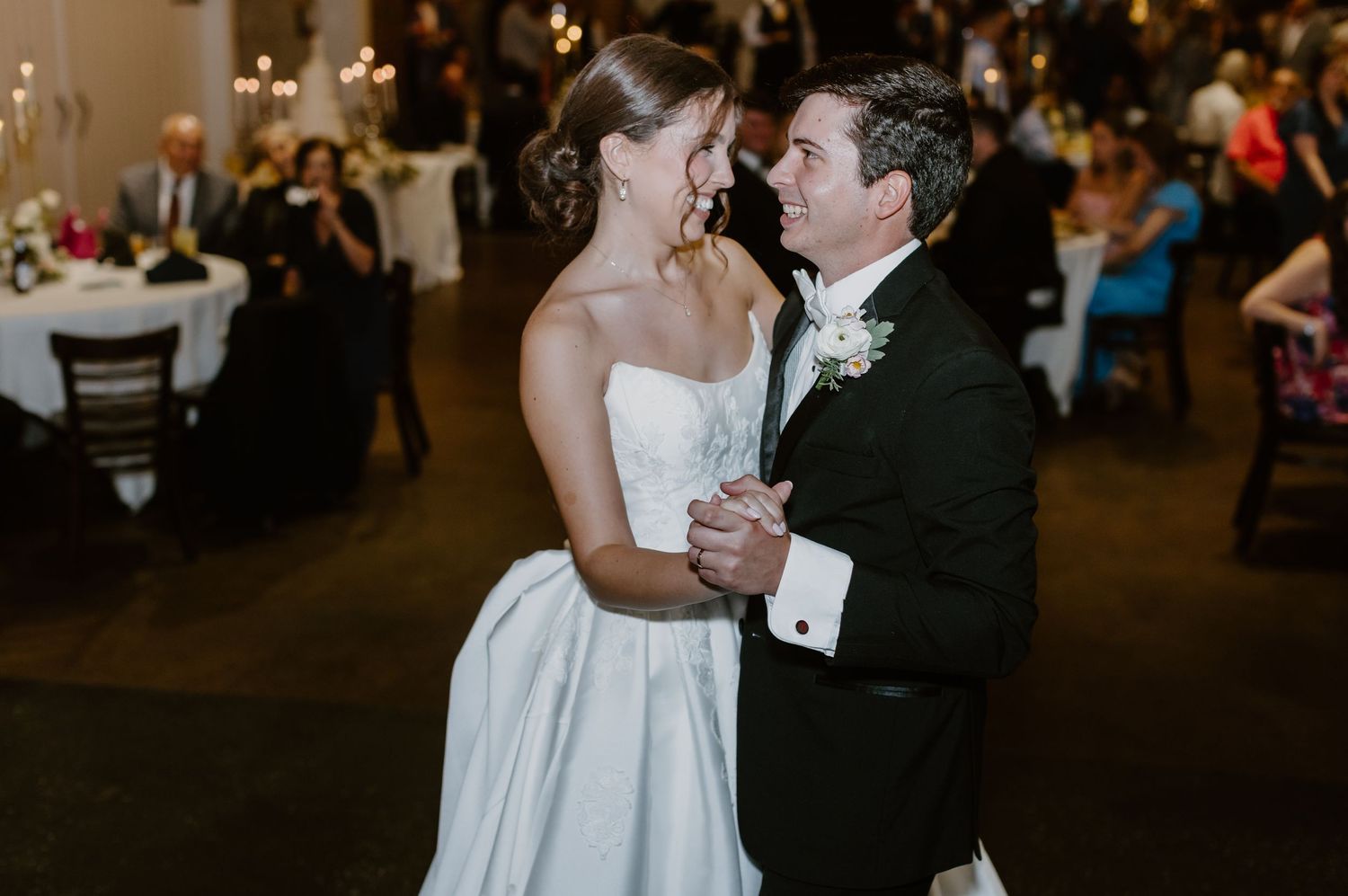 Bride and groom share their first dance at elegant wedding reception, bride in strapless gown, groom in black tuxedo.