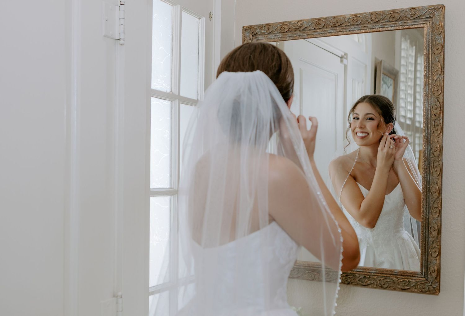 Bride in white gown and veil adjusting earrings while looking in ornate mirror on wedding day.