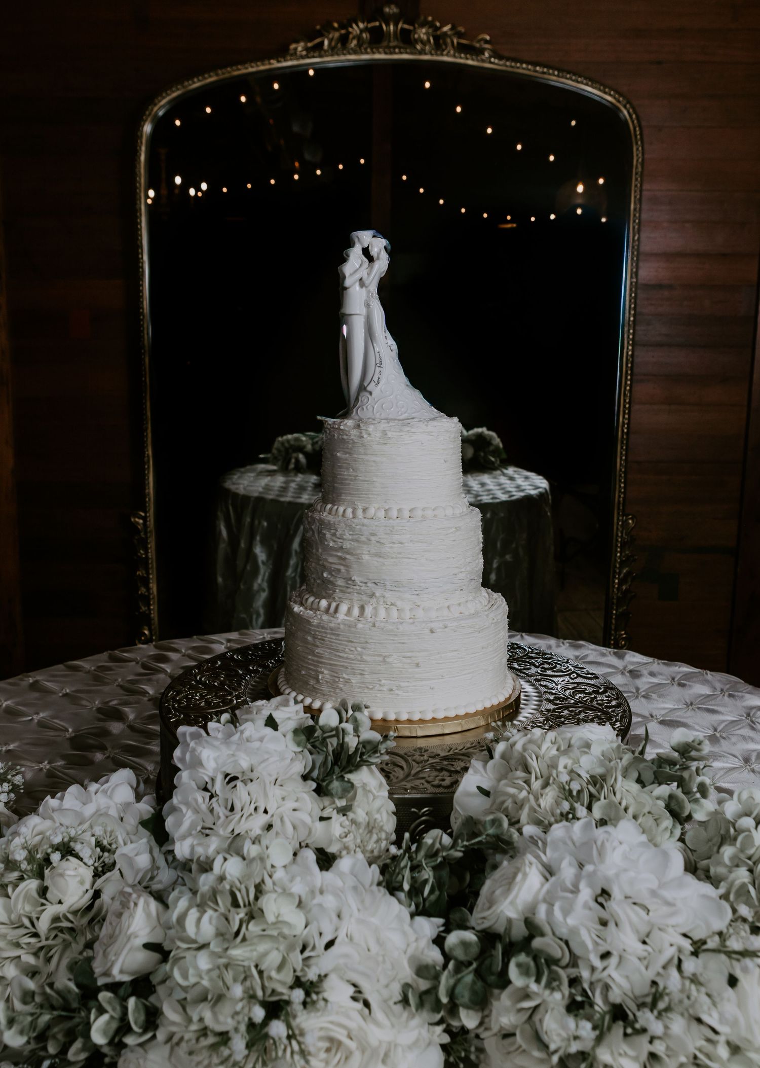 Elegant four-tier white wedding cake with bride and groom topper, surrounded by white flowers, reflected in ornate gold mirror.