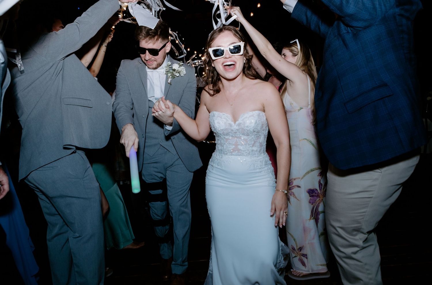 Bride in strapless gown and white sunglasses dancing joyfully with guests at a lively wedding reception celebration.
