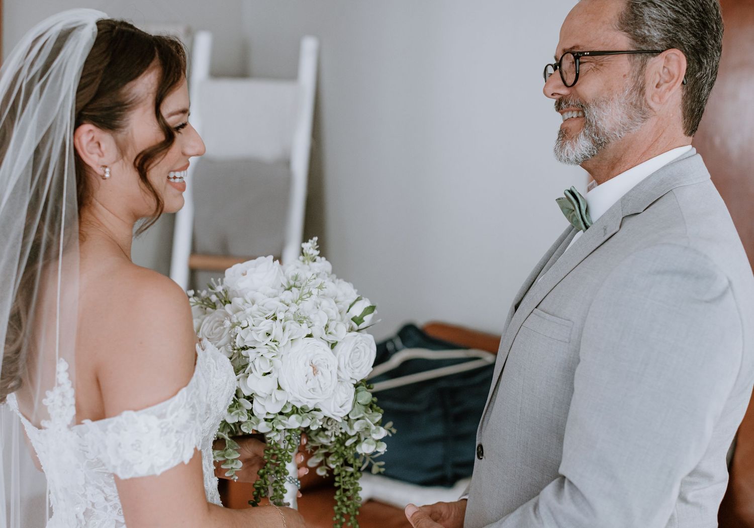 A smiling bride in a white lace gown holds a bouquet while facing an older man in a gray suit at a wedding.