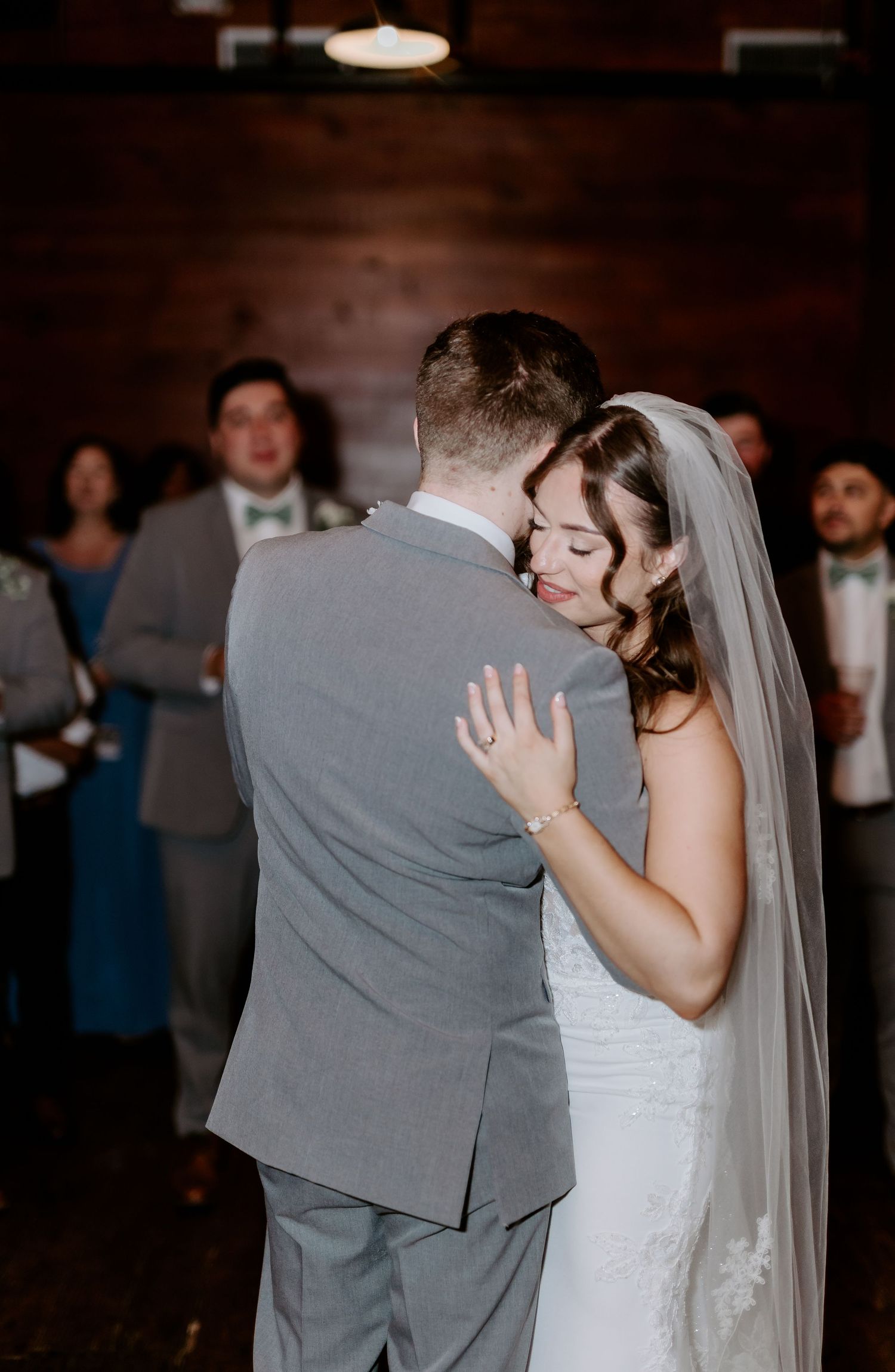 Bride in white lace gown and veil embraces groom during emotional first dance at rustic wedding reception.