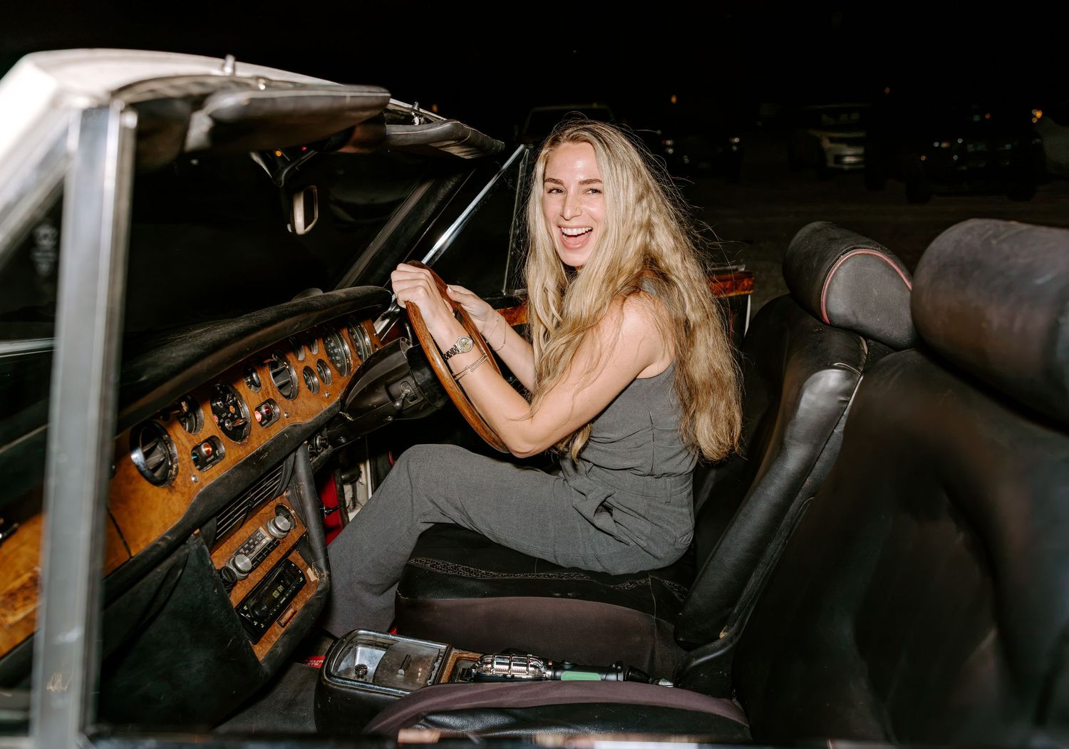 Smiling woman with long blonde hair sits in the driver's seat of a classic convertible car with a wood-paneled dashboard at night.