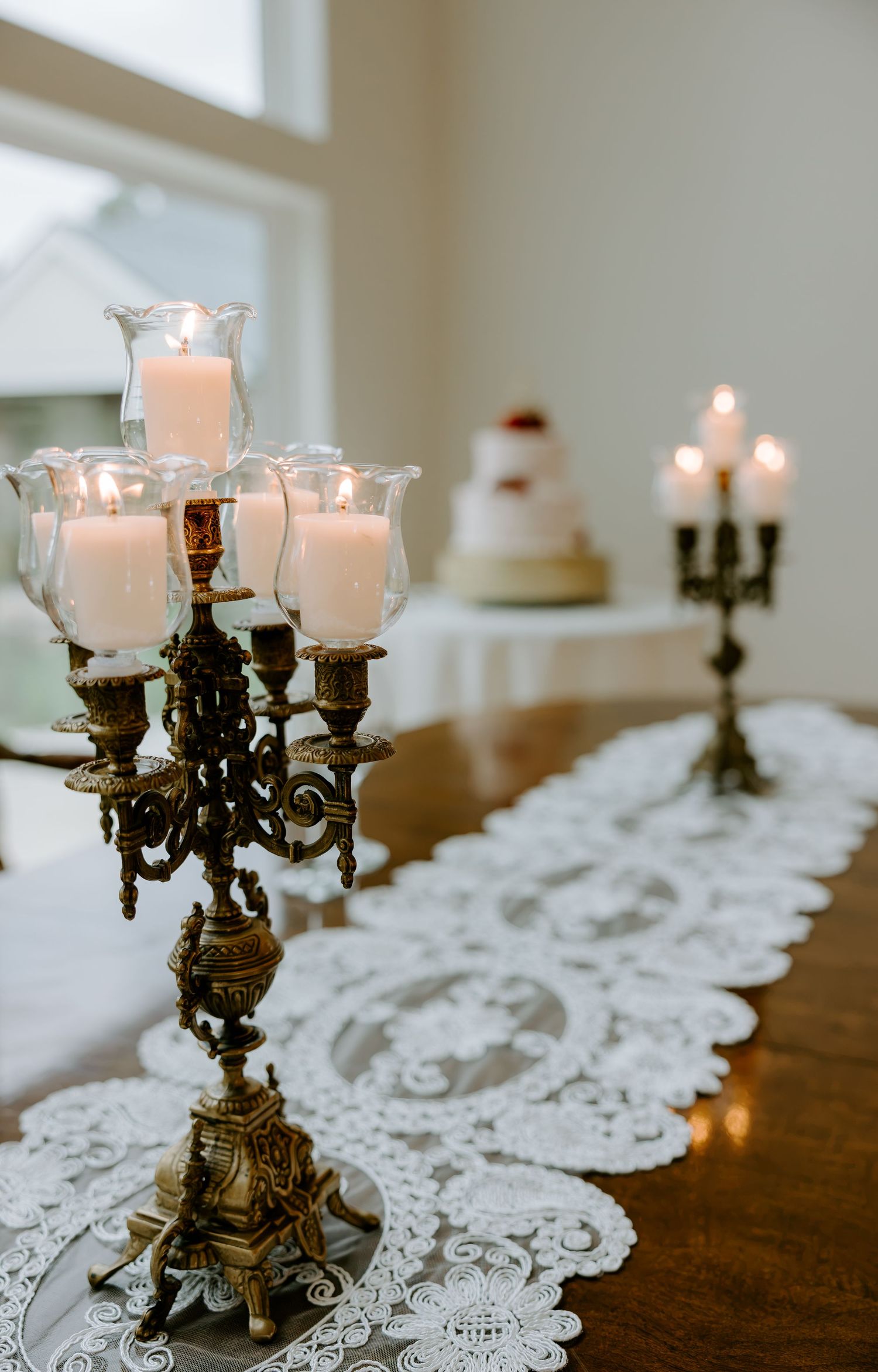Ornate antique brass candelabras with lit white candles in glass holders on a lace table runner, with a wedding cake in the background.