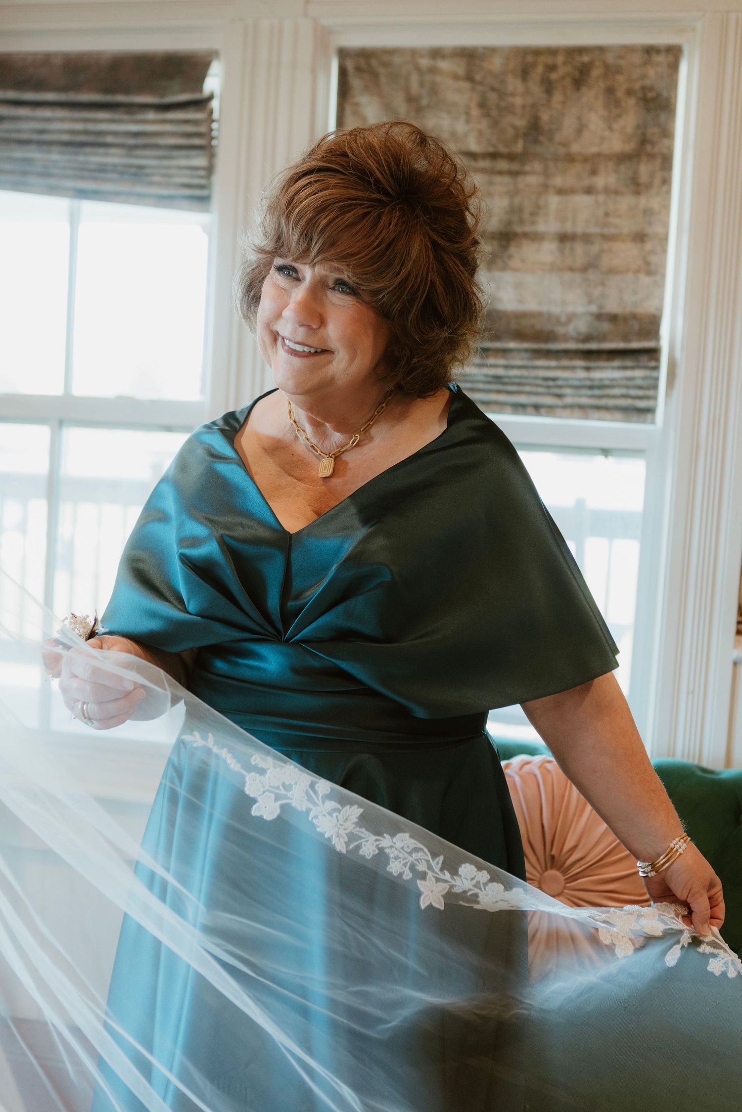 Woman in teal satin dress smiles while holding a lace-trimmed bridal veil near a bright window.