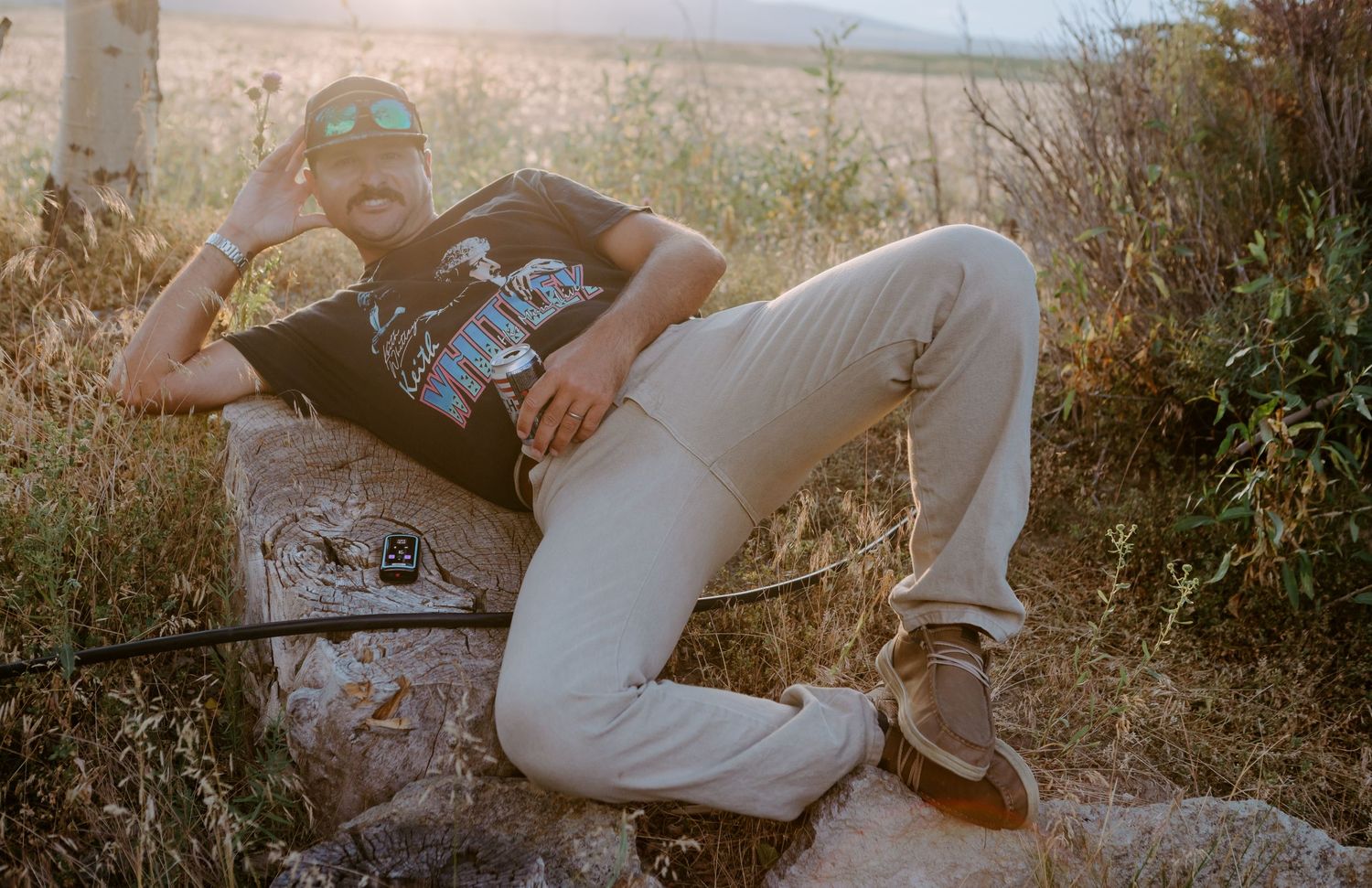 A bearded man relaxes on a rock outdoors, wearing a black graphic tee and light pants, surrounded by tall grass.