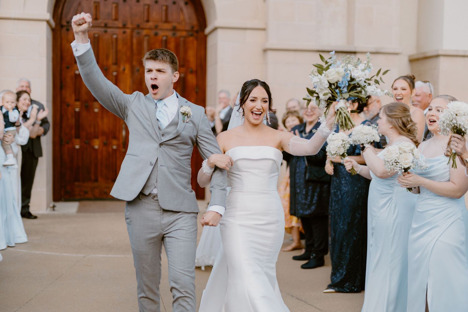A newly married couple celebrates joyfully while exiting their wedding ceremony as guests cheer with blue floral bouquets.