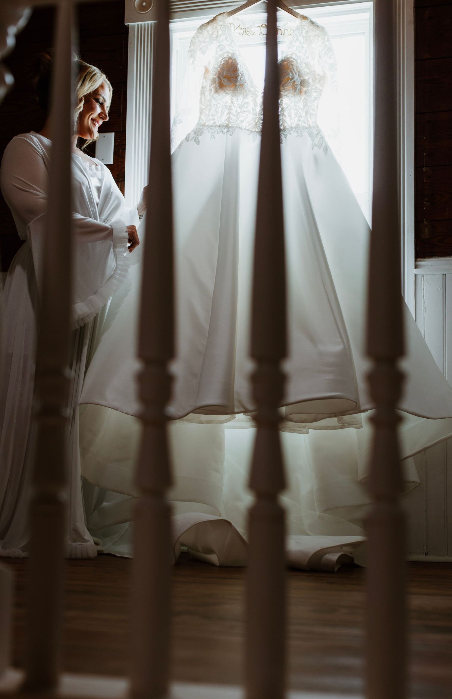 A white wedding dress hangs in a room with elegant banister railings creating a dreamy, romantic atmosphere.