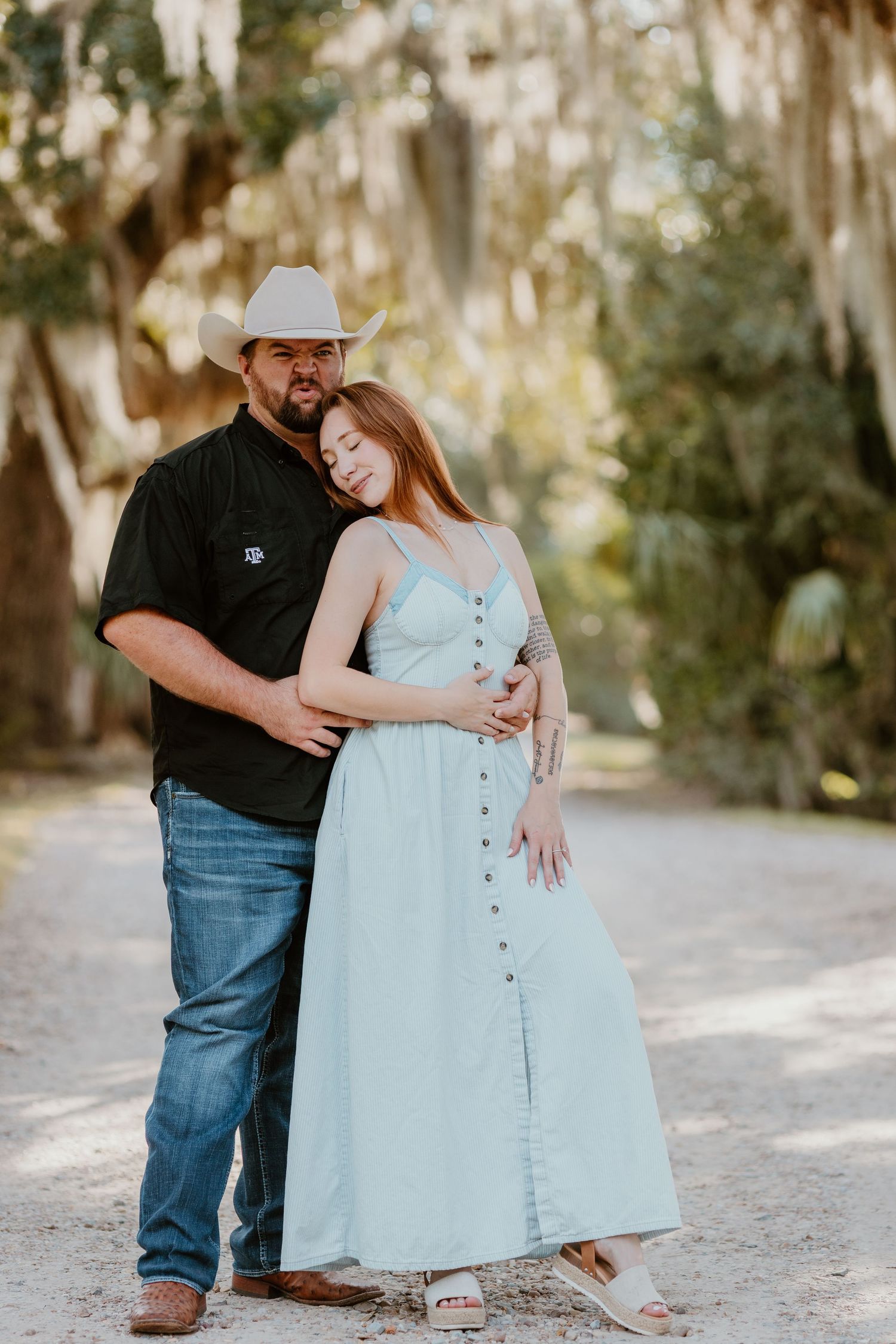 A couple in casual attire embraces on a sunlit path beneath Spanish moss draped trees.