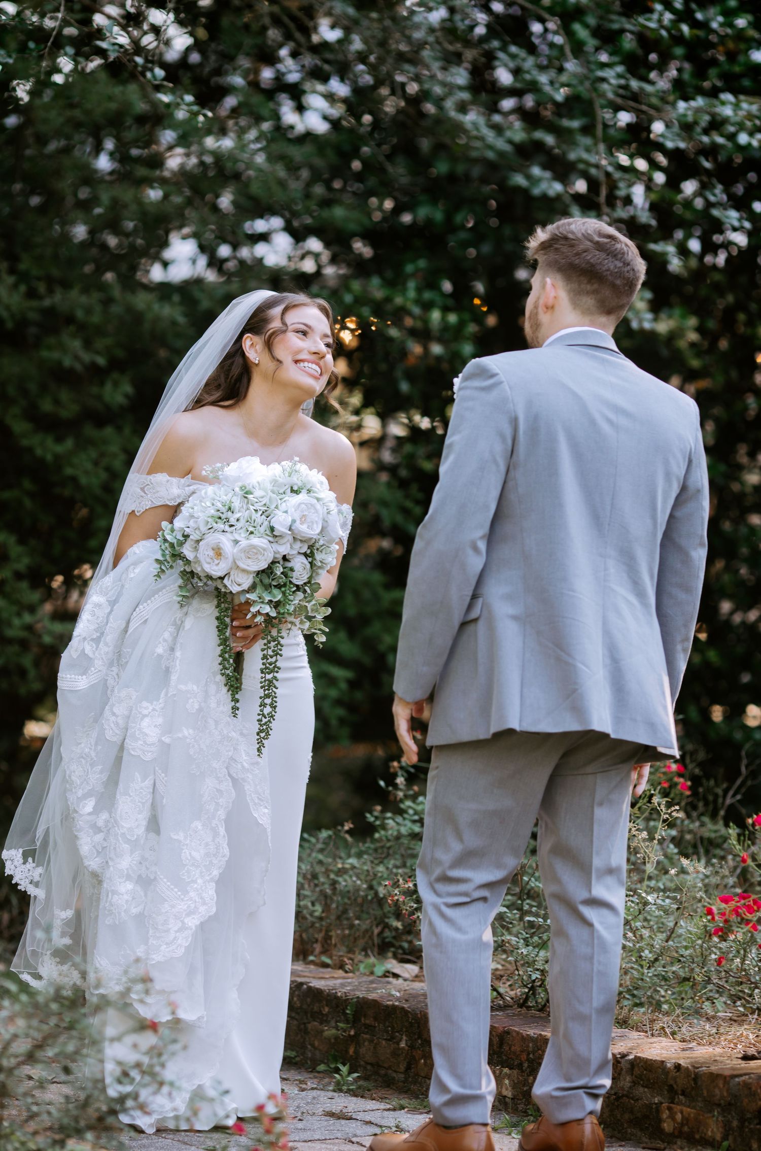Bride in lace gown holding white bouquet smiles at groom in gray suit on stone steps surrounded by lush greenery.