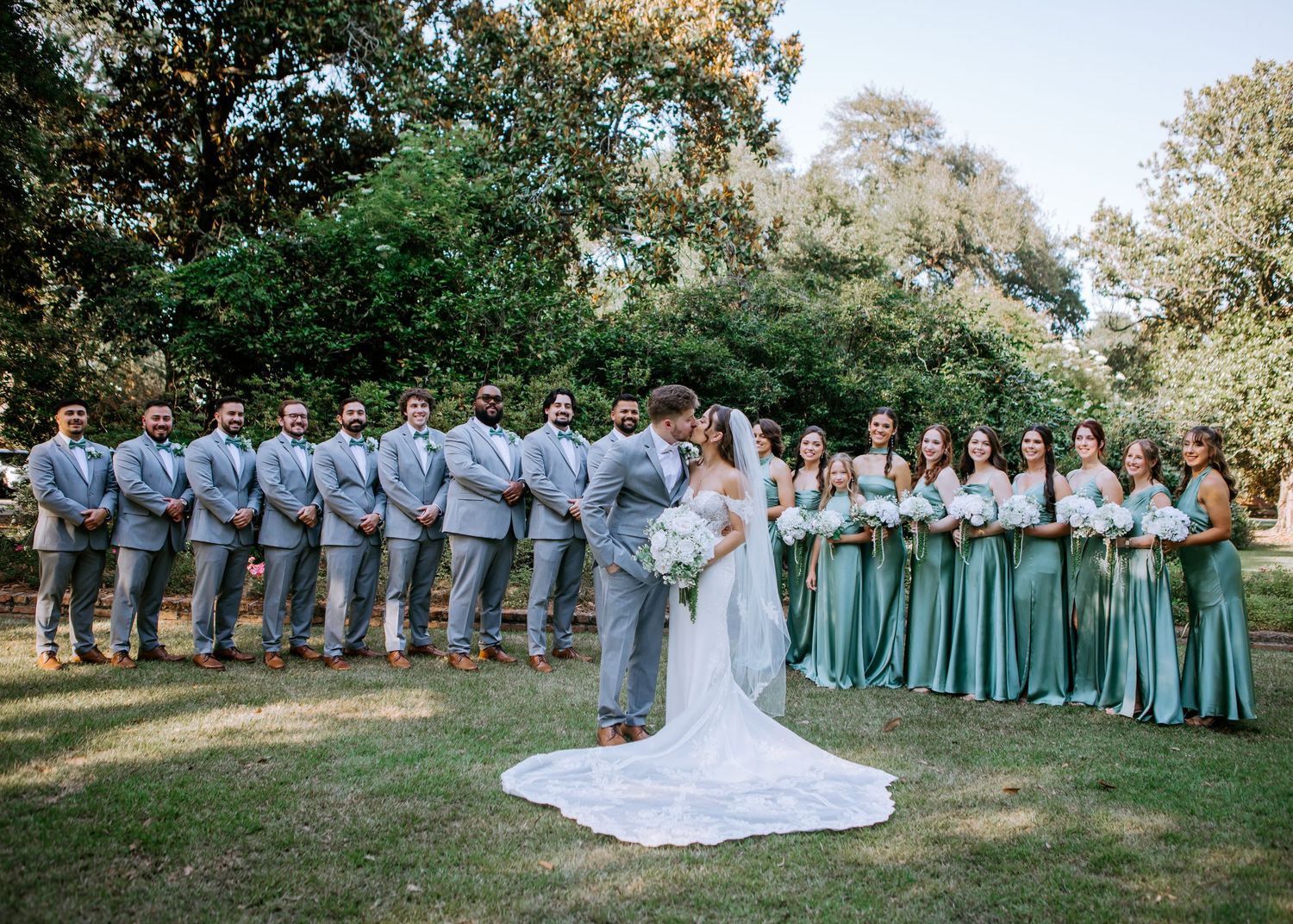 Wedding party poses outdoors, bride and groom kissing flanked by groomsmen in gray suits and bridesmaids in sage green gowns.