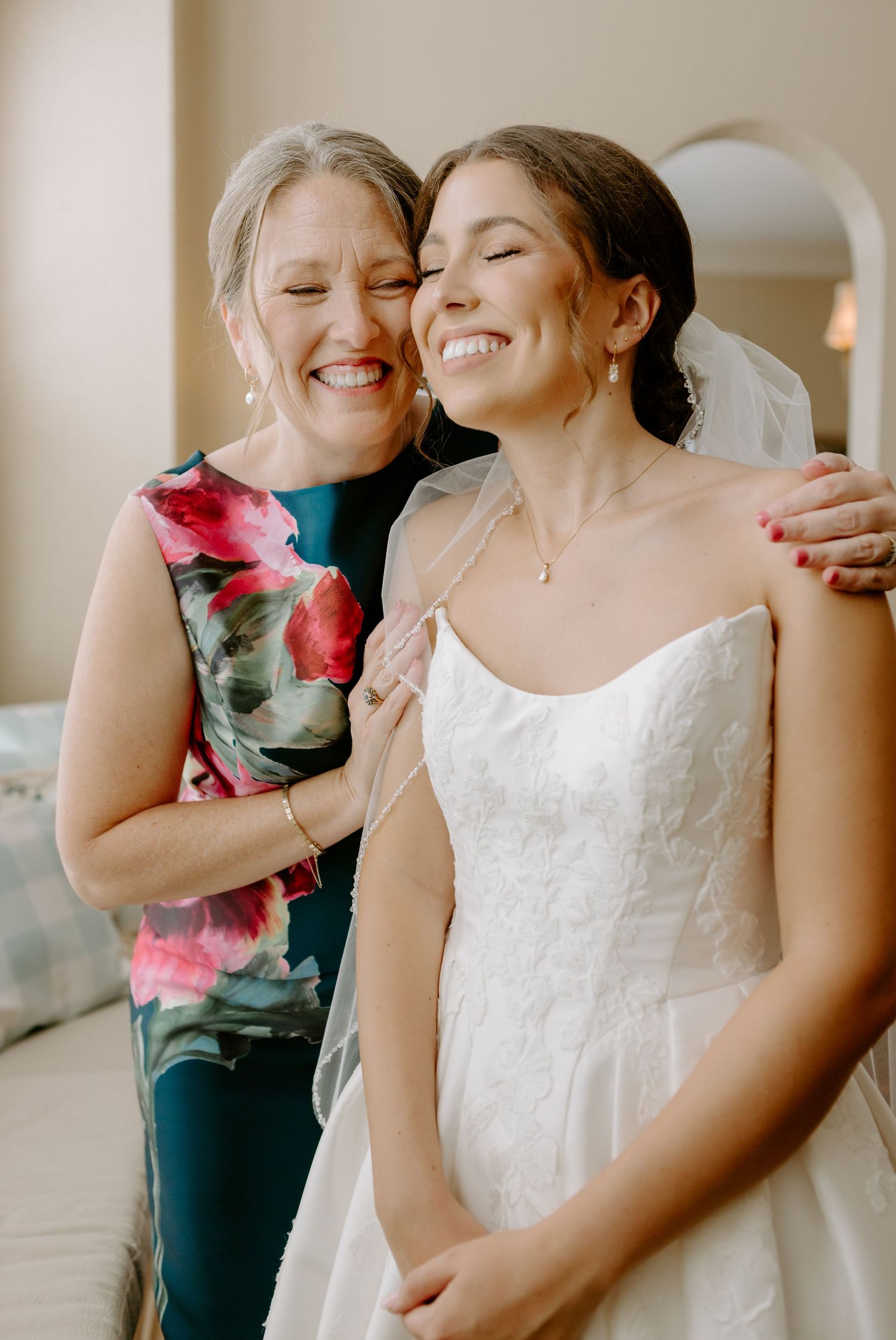 Joyful bride in strapless lace gown shares a happy embrace with older woman in floral dress before wedding ceremony.