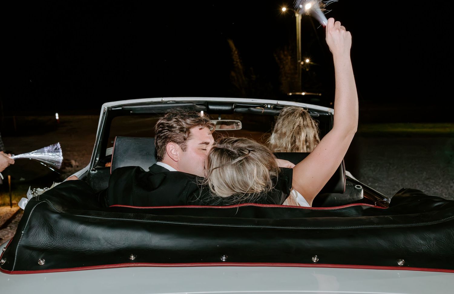 Couple kissing in the back of a black convertible at night while a guest raises a sparkler in the air during a wedding send-off.