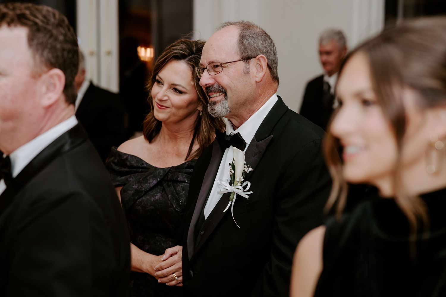 Smiling man in a black tuxedo with a white floral boutonniere stands beside a woman in a black dress at an elegant formal event.